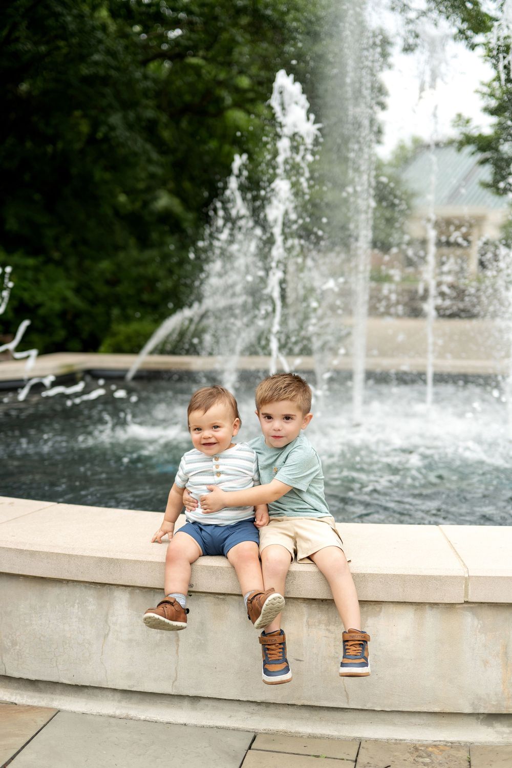 Two young boys sitting on a fountain ledge outdoors, smiling together with water spraying behind them.