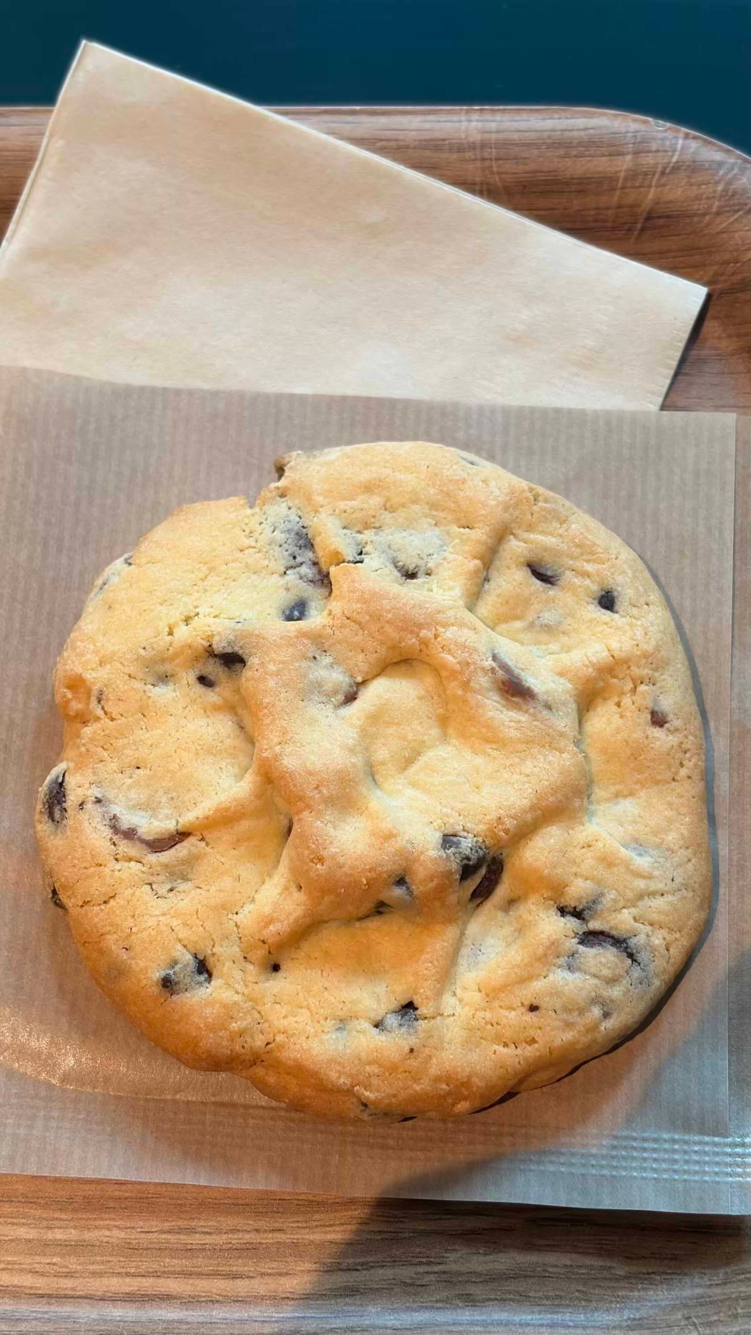 A large golden-brown chocolate chip cookie resting on parchment paper on a wooden tray.