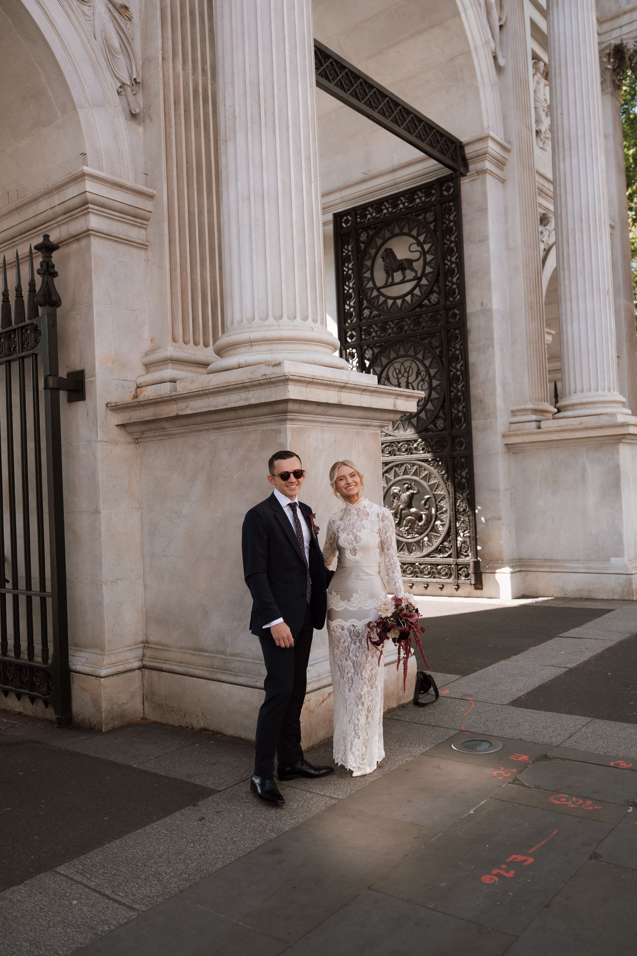 Elegant bride and groom posing in front of ornate iron gates and white marble columns on a city street.