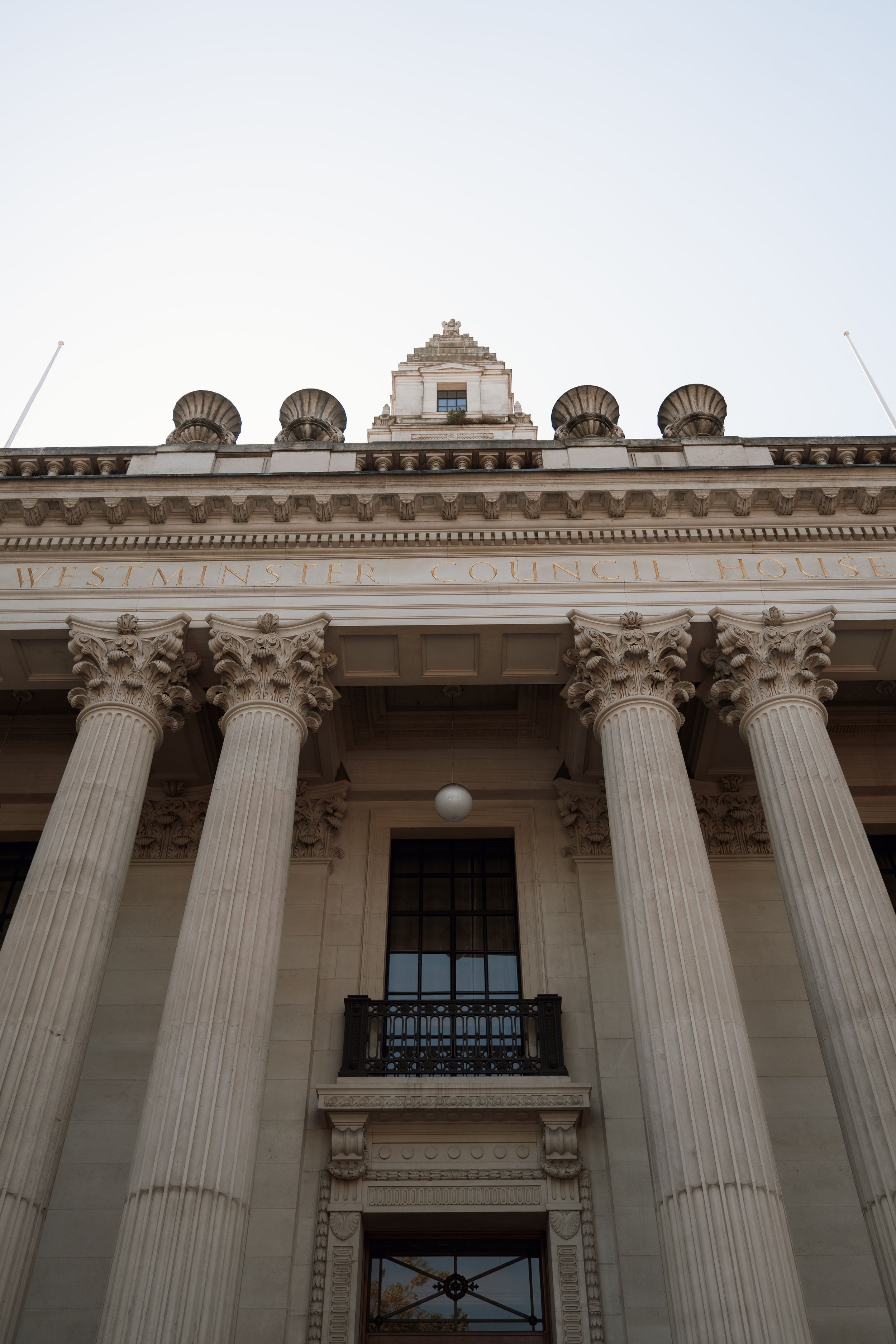 Neoclassical building facade with tall Corinthian columns, ornate cornice, and decorative rooftop elements against a pale sky.