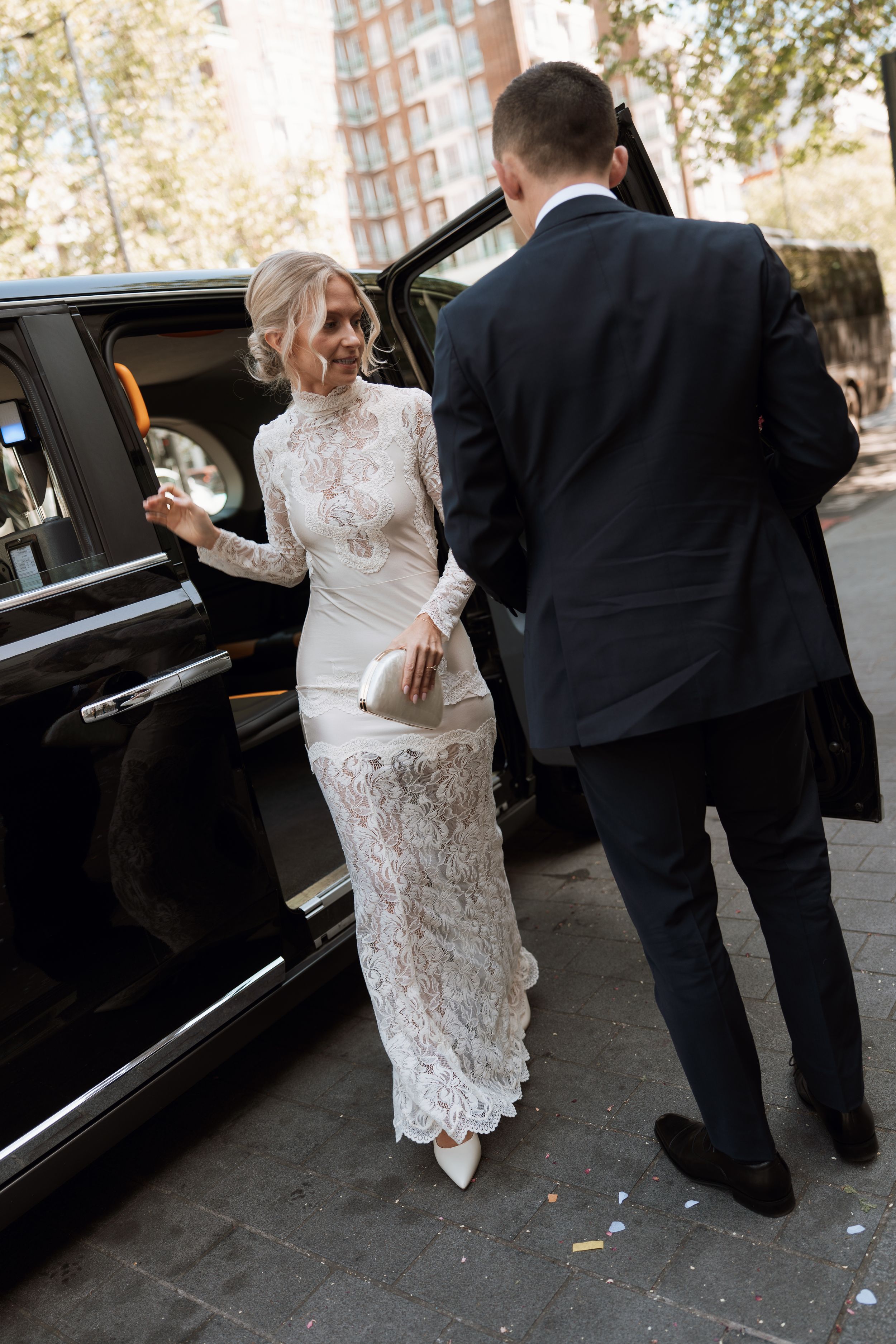 Bride in white lace gown steps out of black luxury car assisted by groom in dark suit on city street.