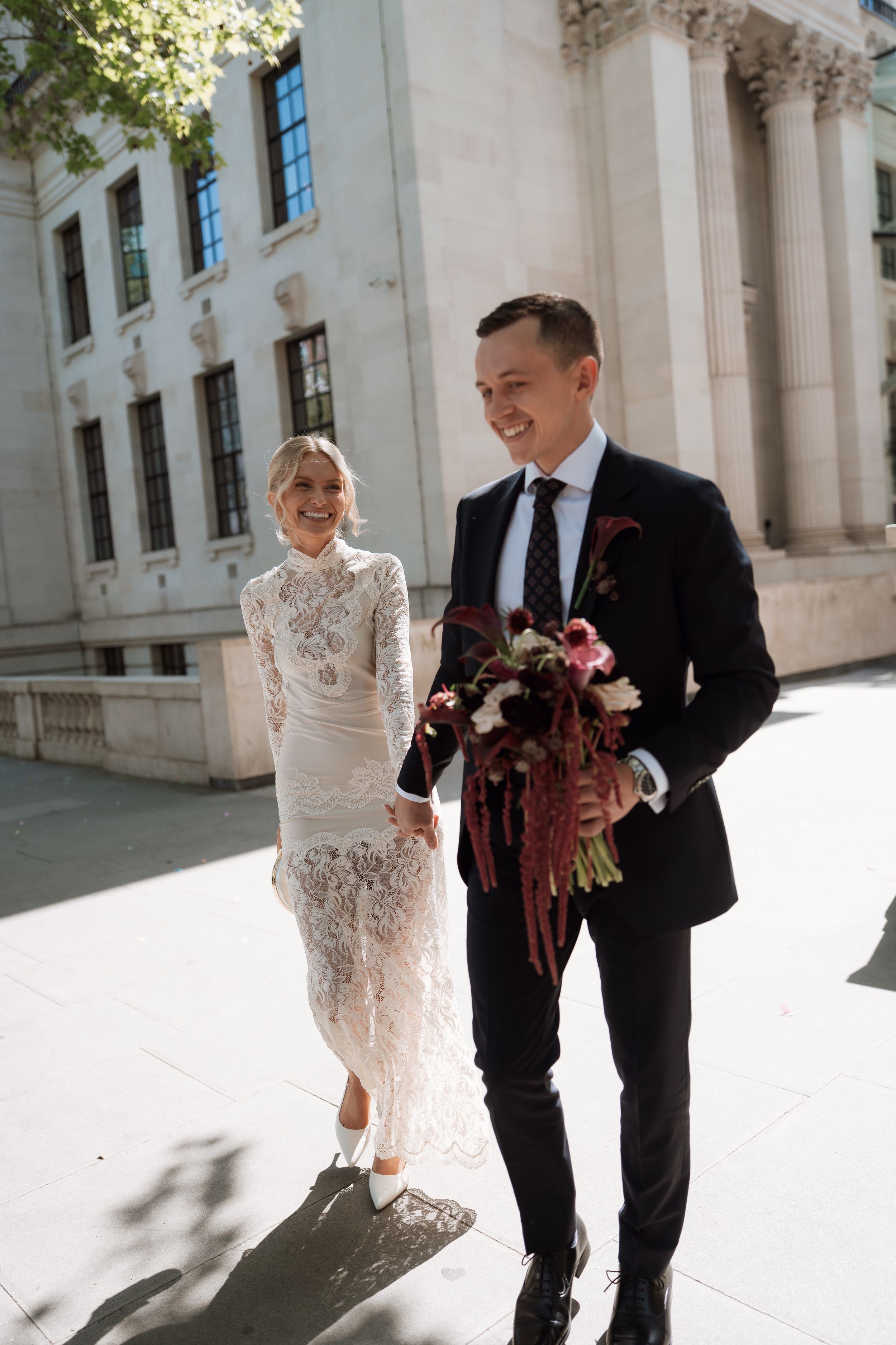 Bride in lace dress and groom in black suit walk together holding a bouquet outside a grand building.