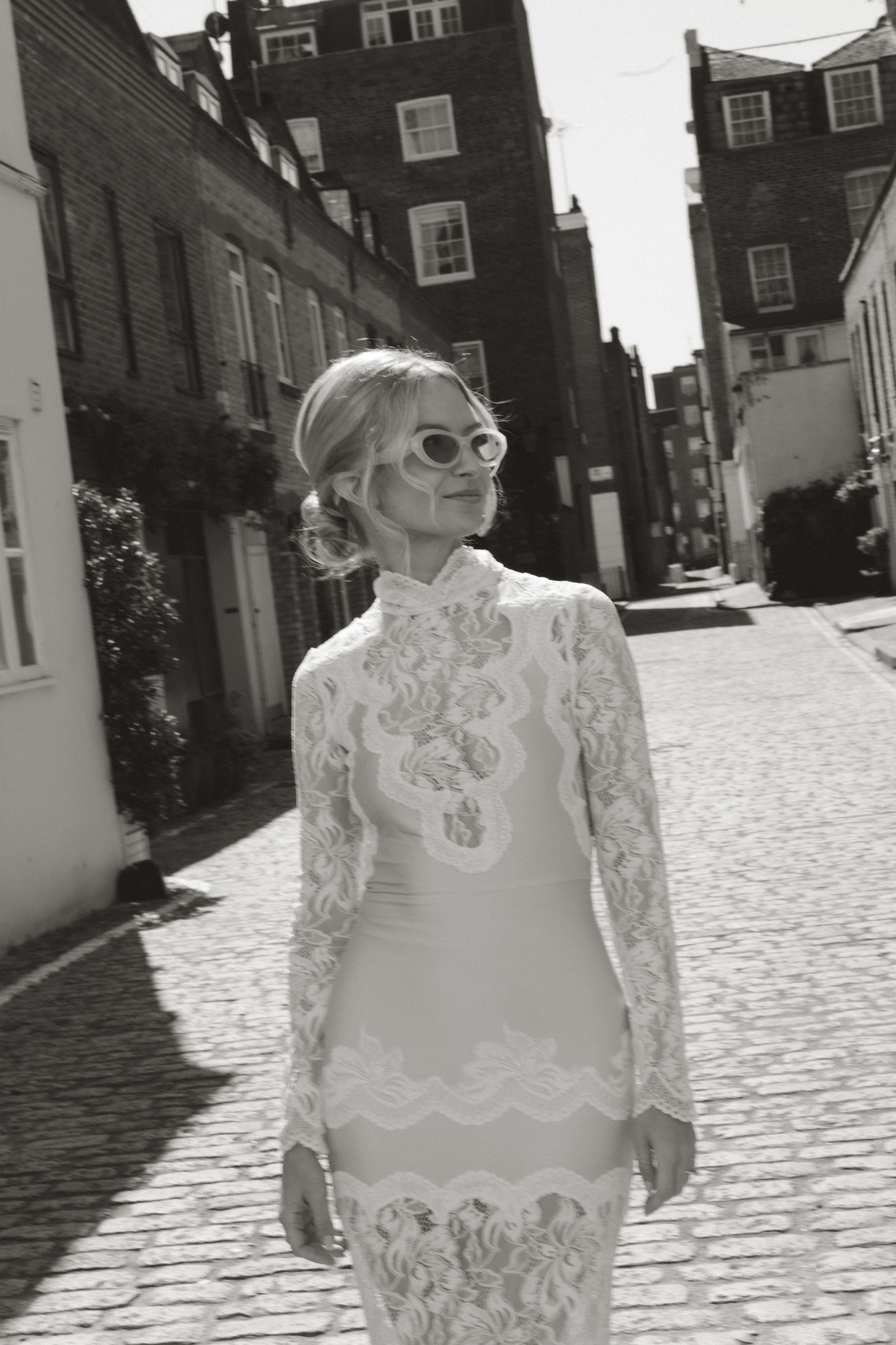 Woman in lace long-sleeve dress and sunglasses posing on cobblestone street in black and white photo.