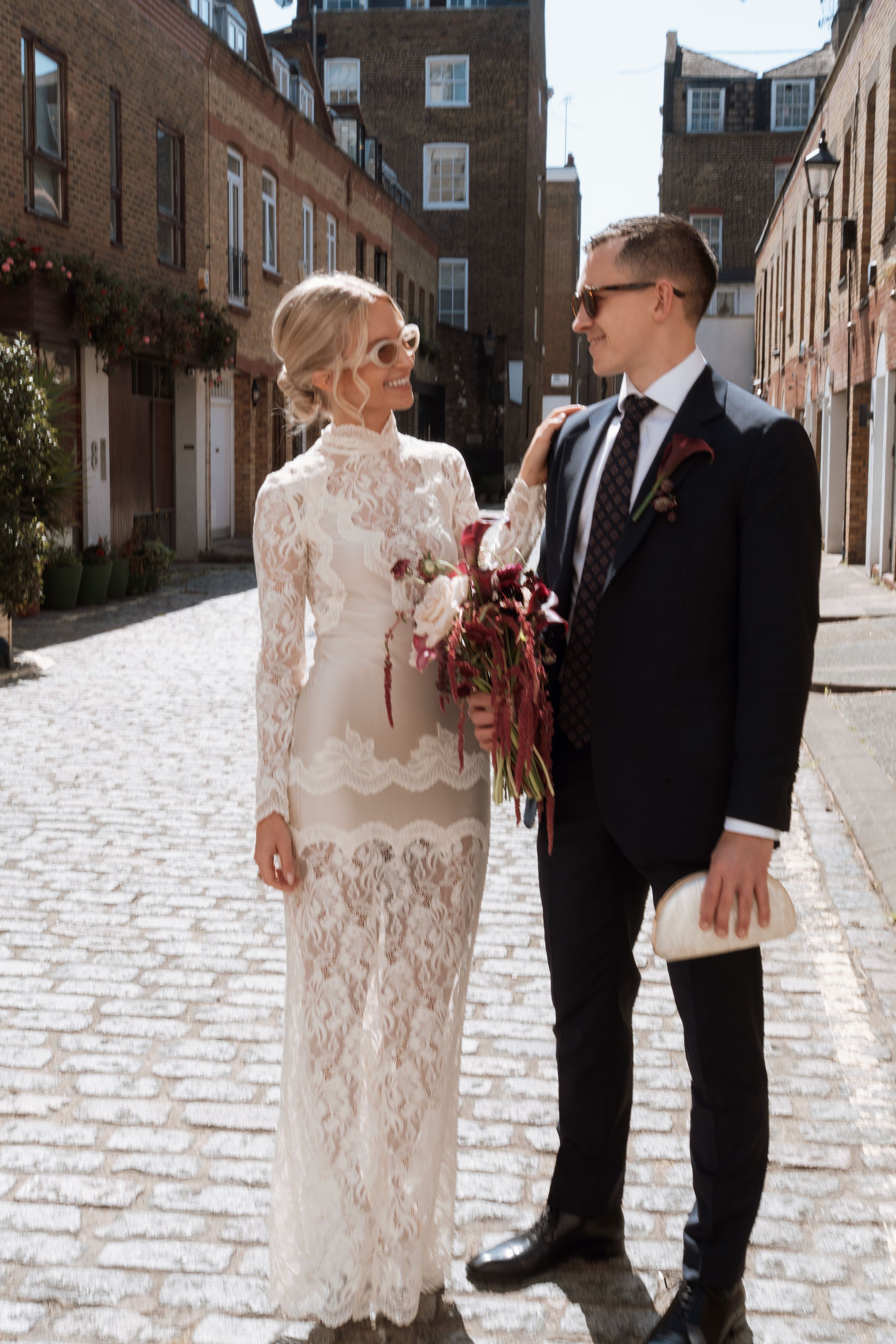 Bride in white lace gown and groom in black suit pose on cobblestone street holding burgundy bouquet.