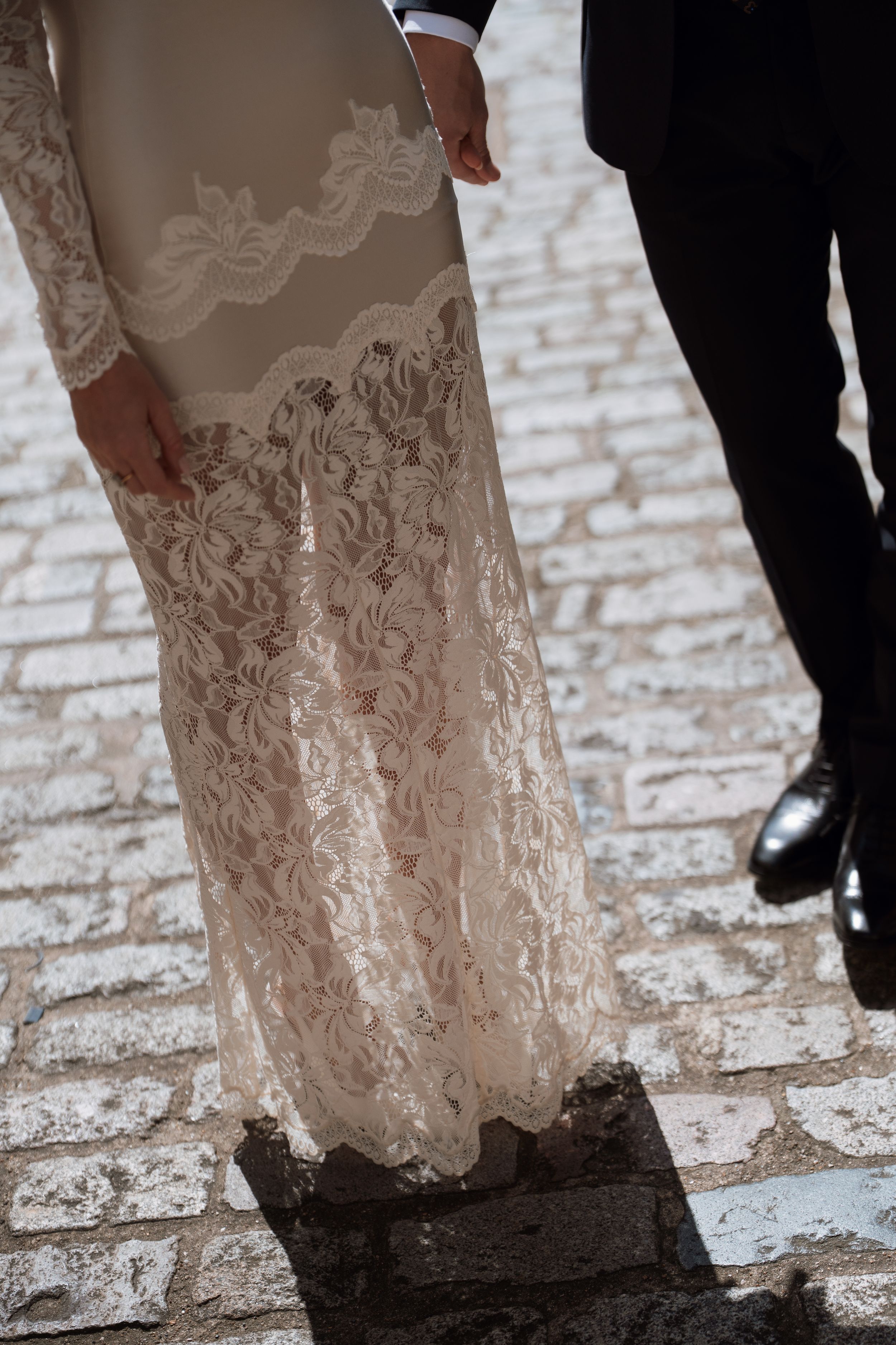 Bride in elegant long-sleeve lace wedding gown standing on cobblestone with groom in black suit beside her.