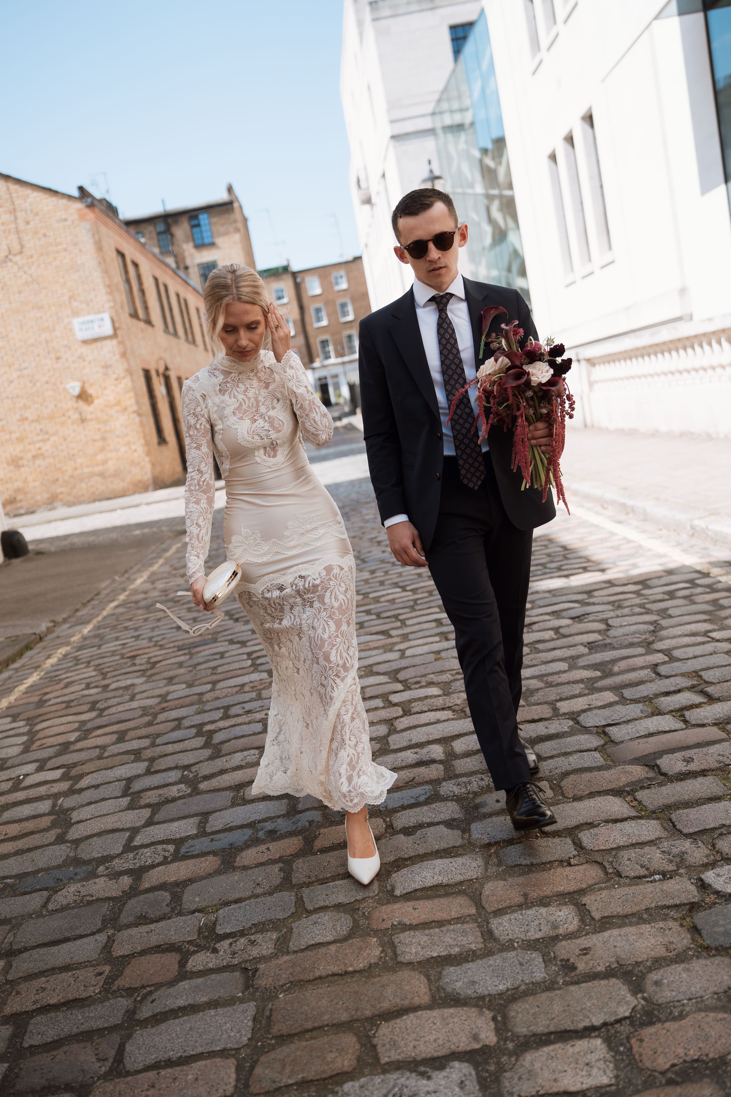 Bride in lace gown and groom in black suit walk cobblestone street holding bouquet on wedding day.