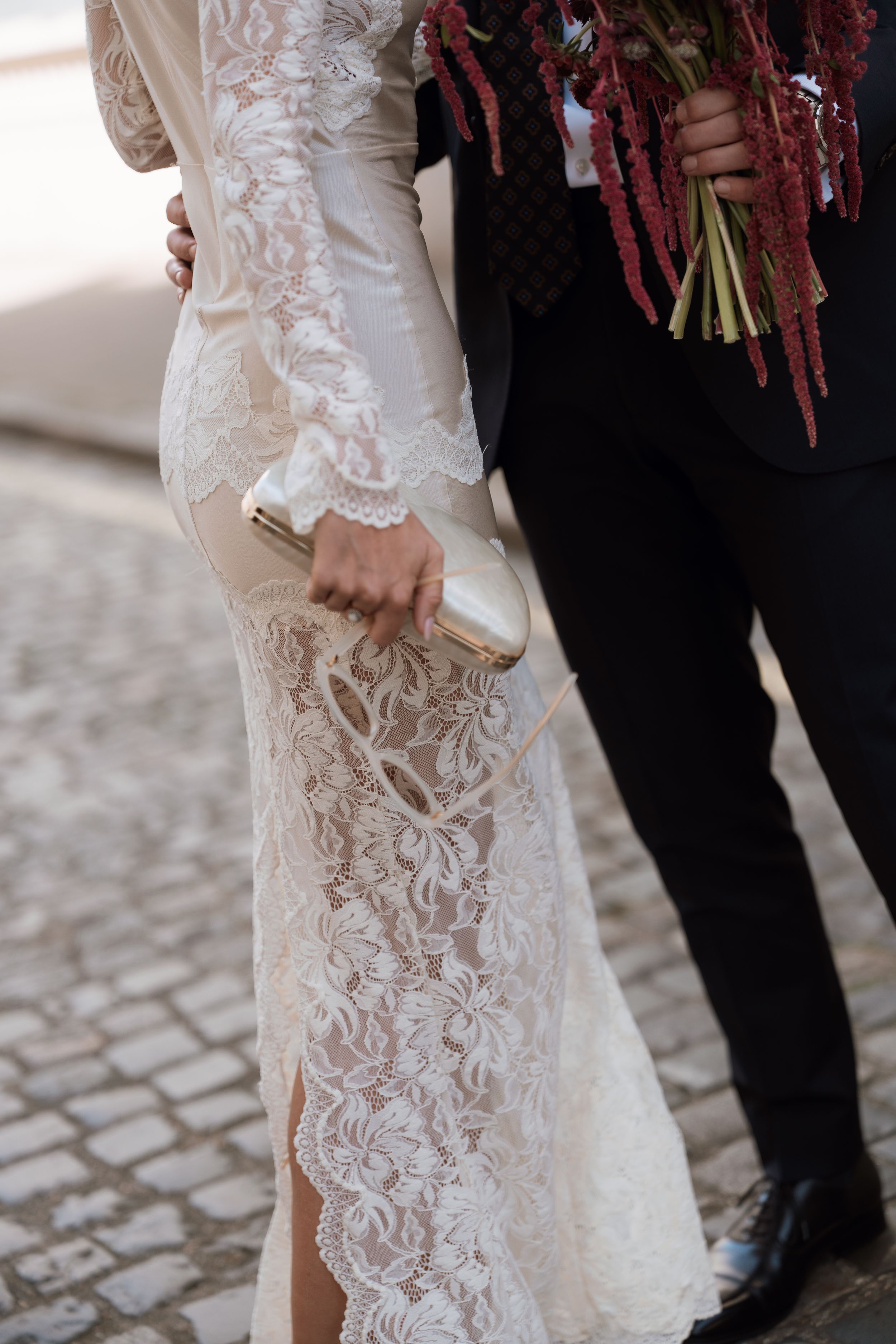 Bride in elegant lace wedding dress holding a clutch, standing beside groom with red floral bouquet on cobblestone.