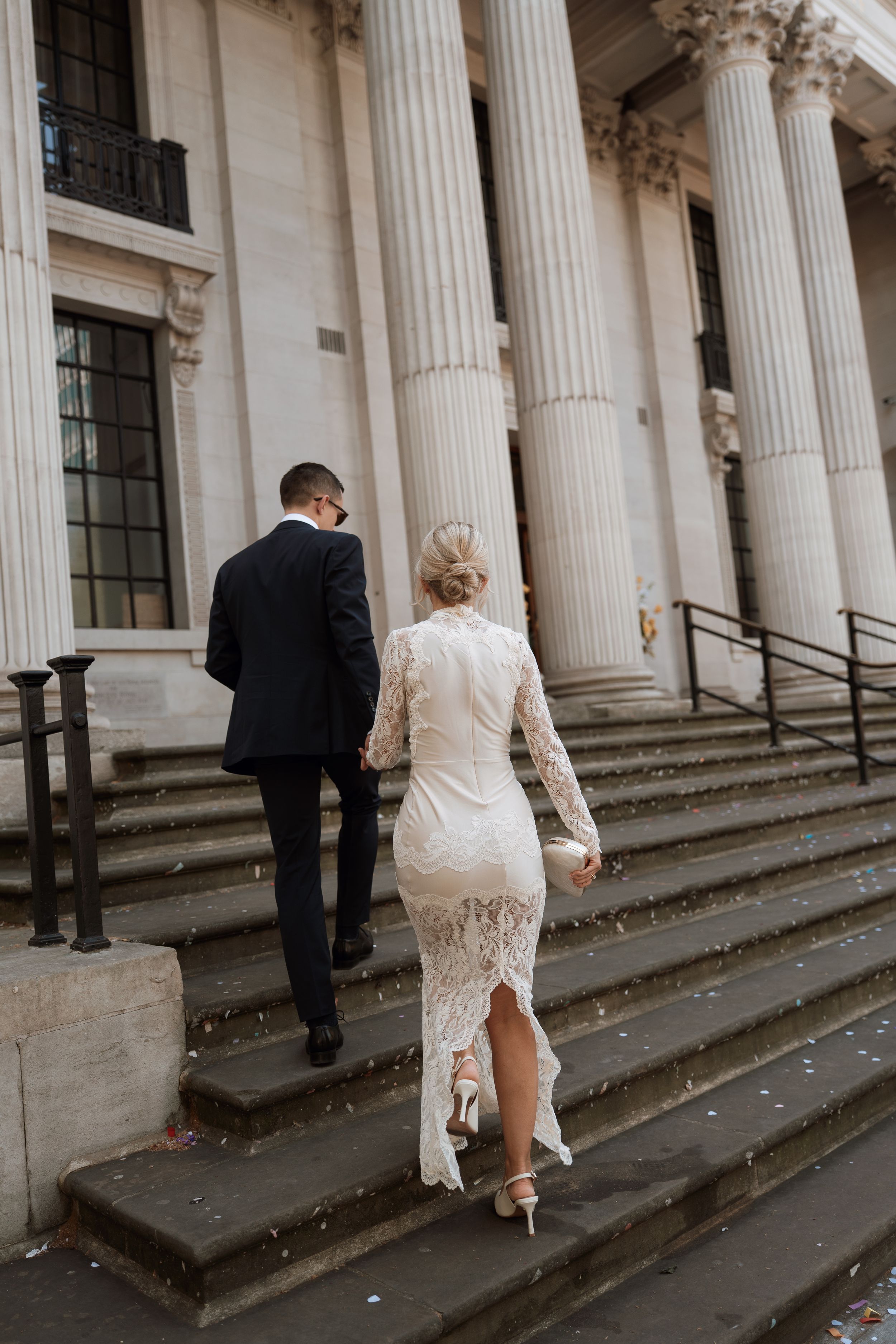 Bride in white lace dress and groom in dark suit walk up stone steps of elegant building with columns.