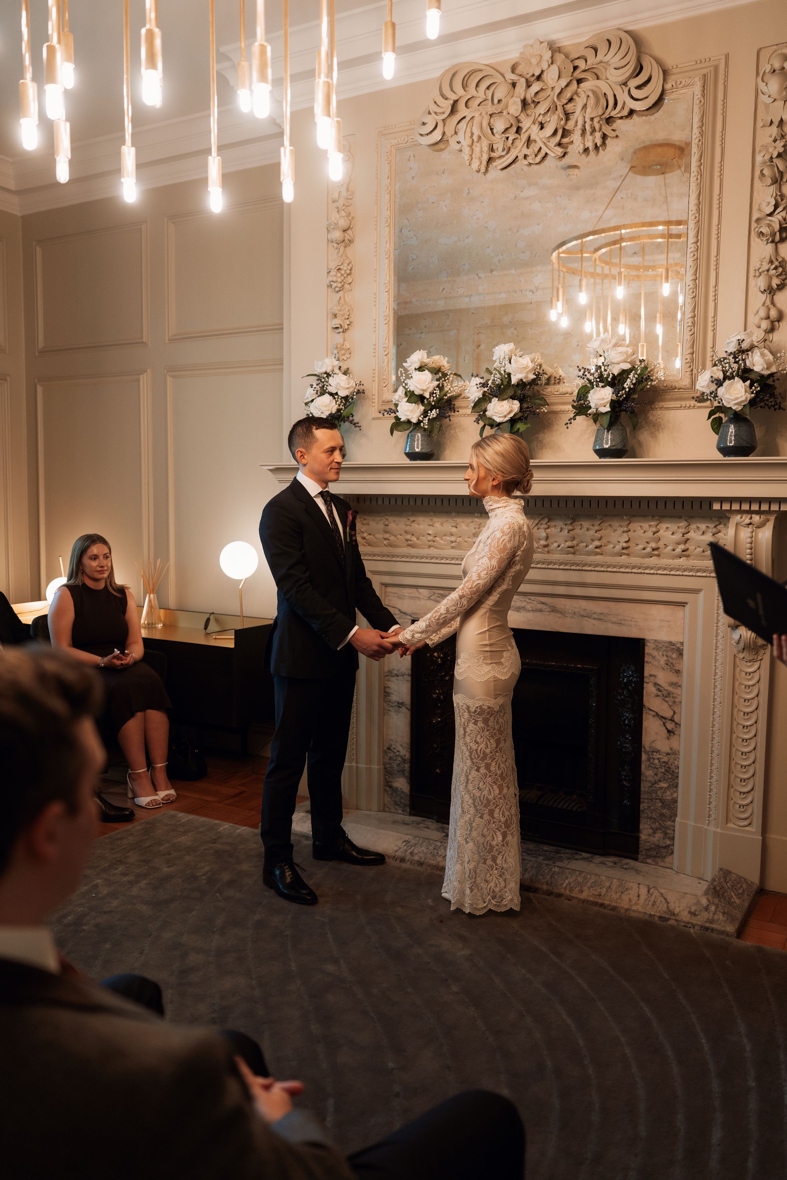 Couple exchanging vows at elegant indoor wedding ceremony by ornate fireplace with white floral arrangements.