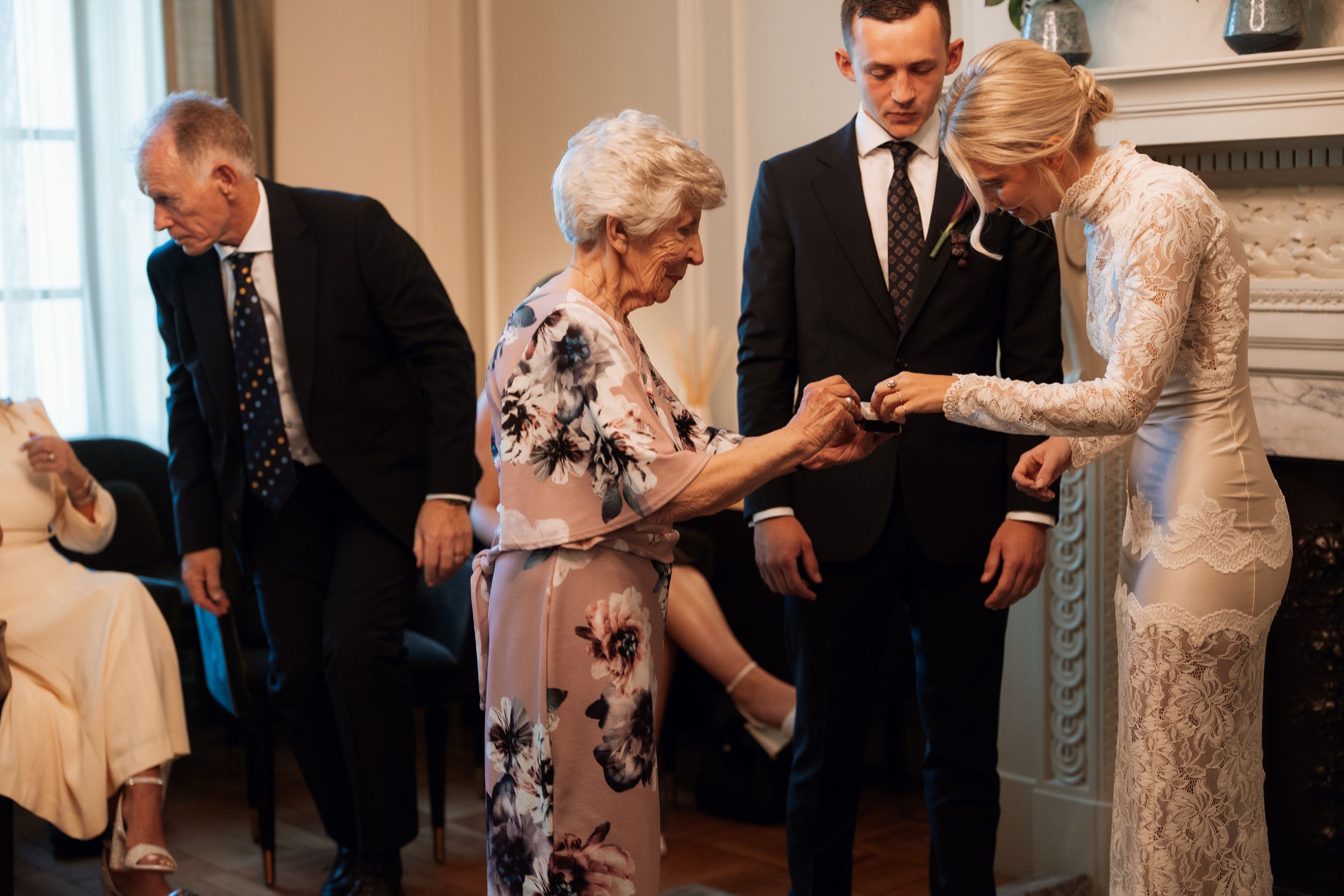 Elderly woman receiving a gift from a blonde woman in lace dress at a formal indoor gathering with guests.