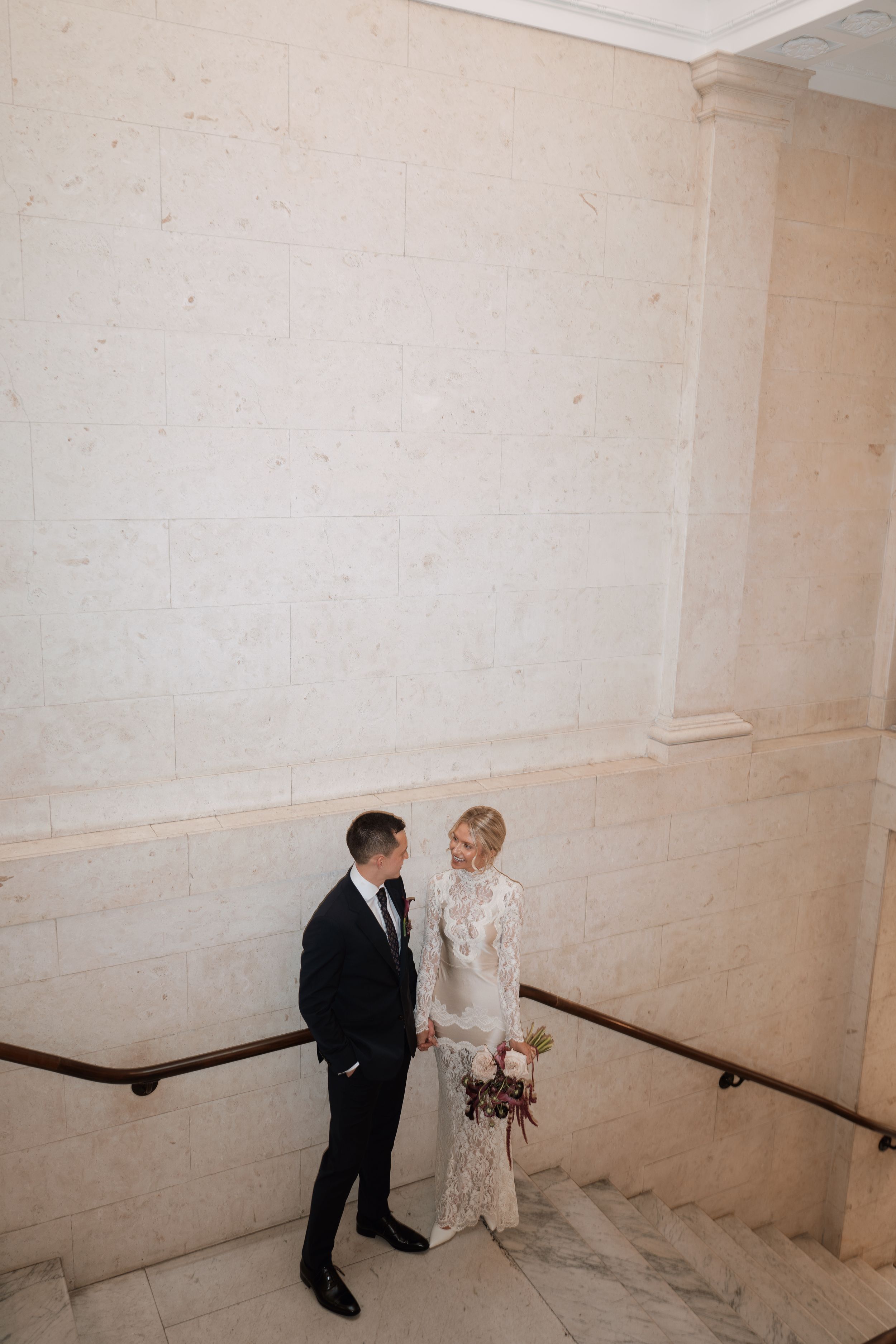 Bride and groom stand on marble staircase, she in lace gown holding bouquet, he in black suit, gazing at each other.