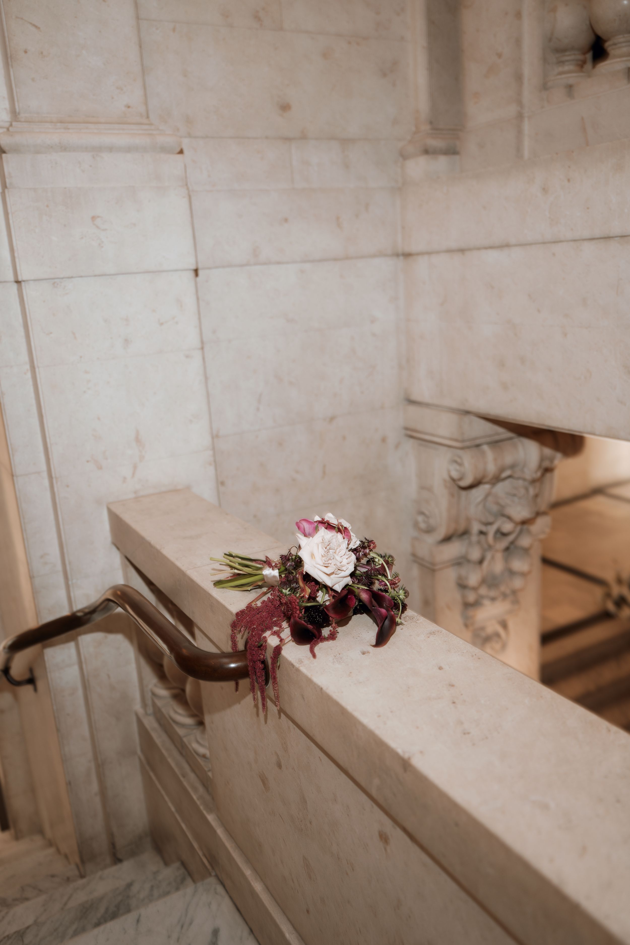 A bridal bouquet with white roses and burgundy ribbons resting on a marble staircase railing in an elegant venue.