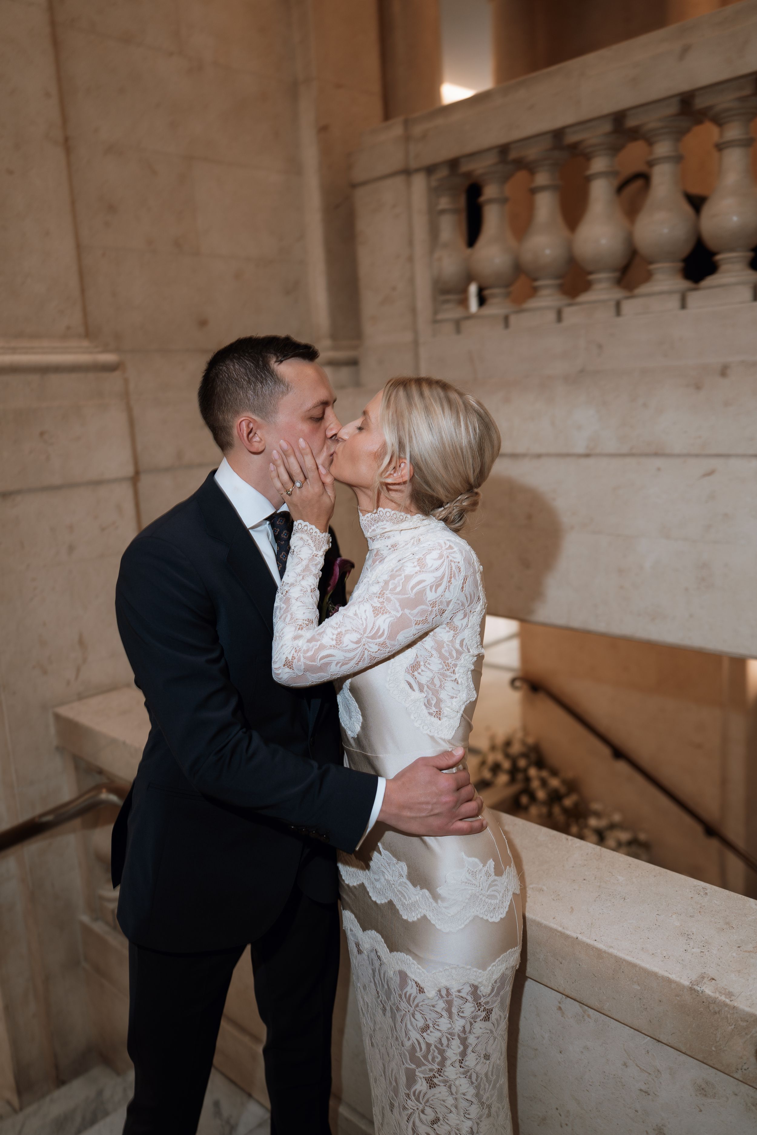 Bride and groom share a romantic kiss on marble staircase, she in lace gown, he in dark suit.