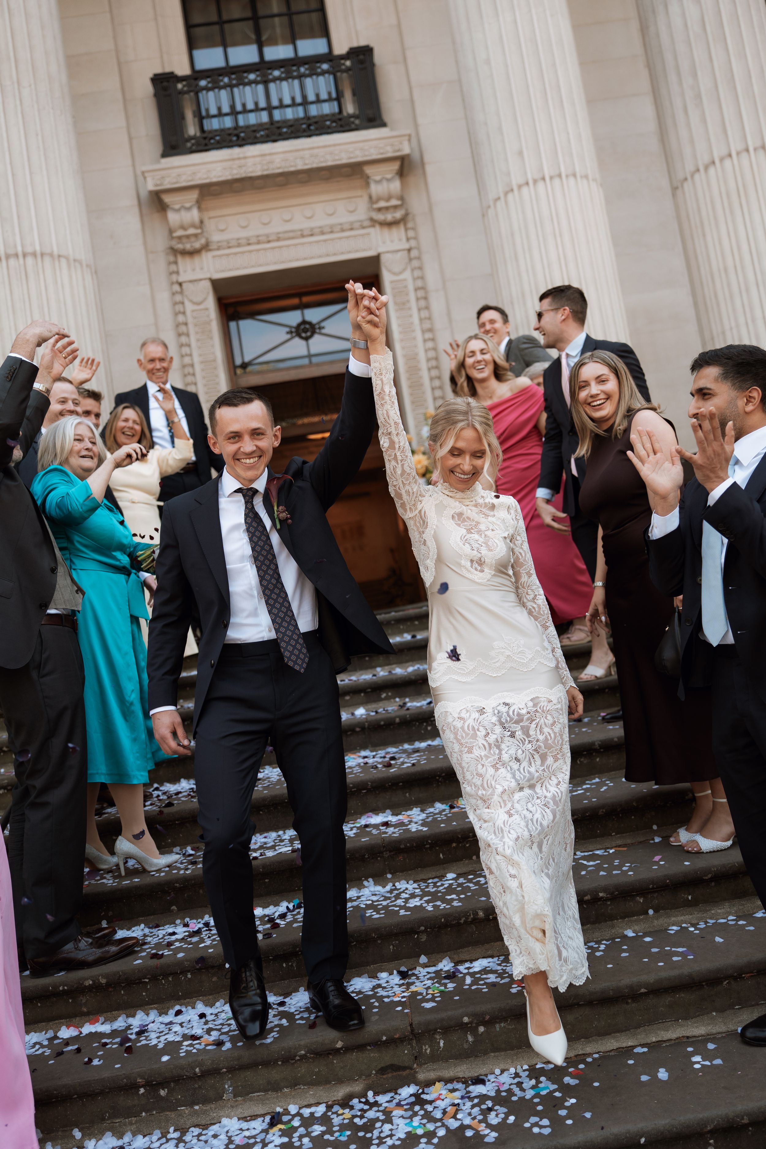 Joyful newlyweds raise hands on church steps surrounded by guests throwing confetti at elegant wedding celebration.