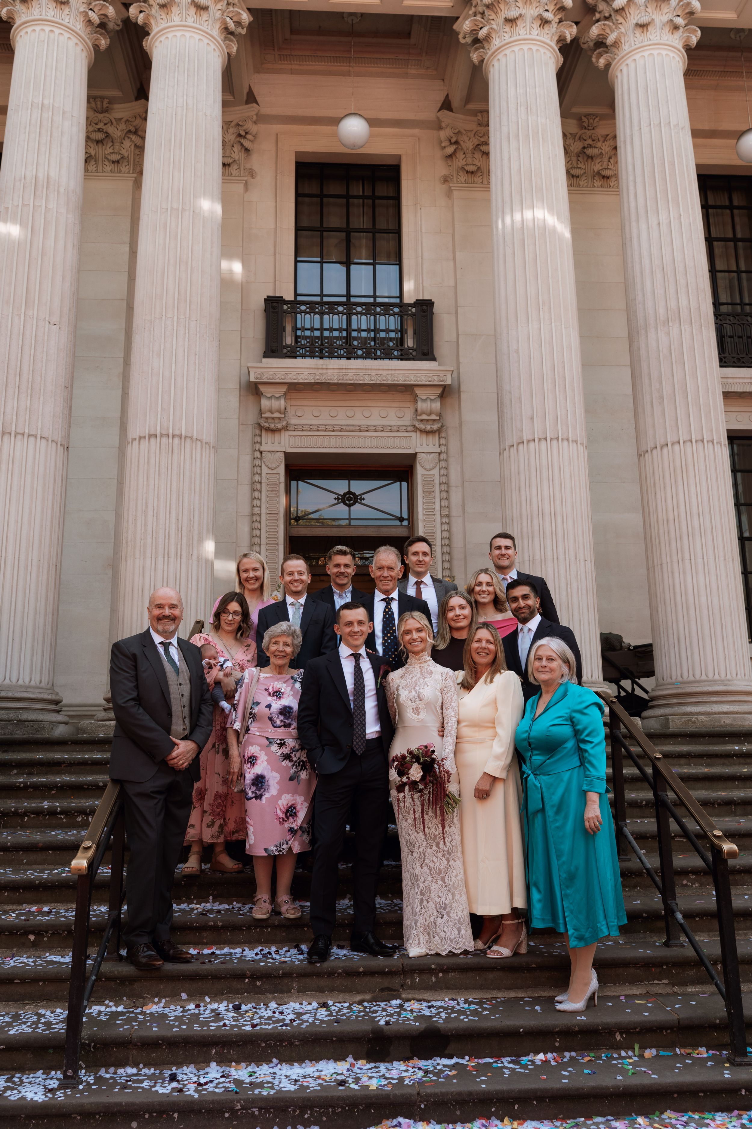 Wedding party posing on grand building steps with columns, confetti falling, bride in white gown with bouquet.