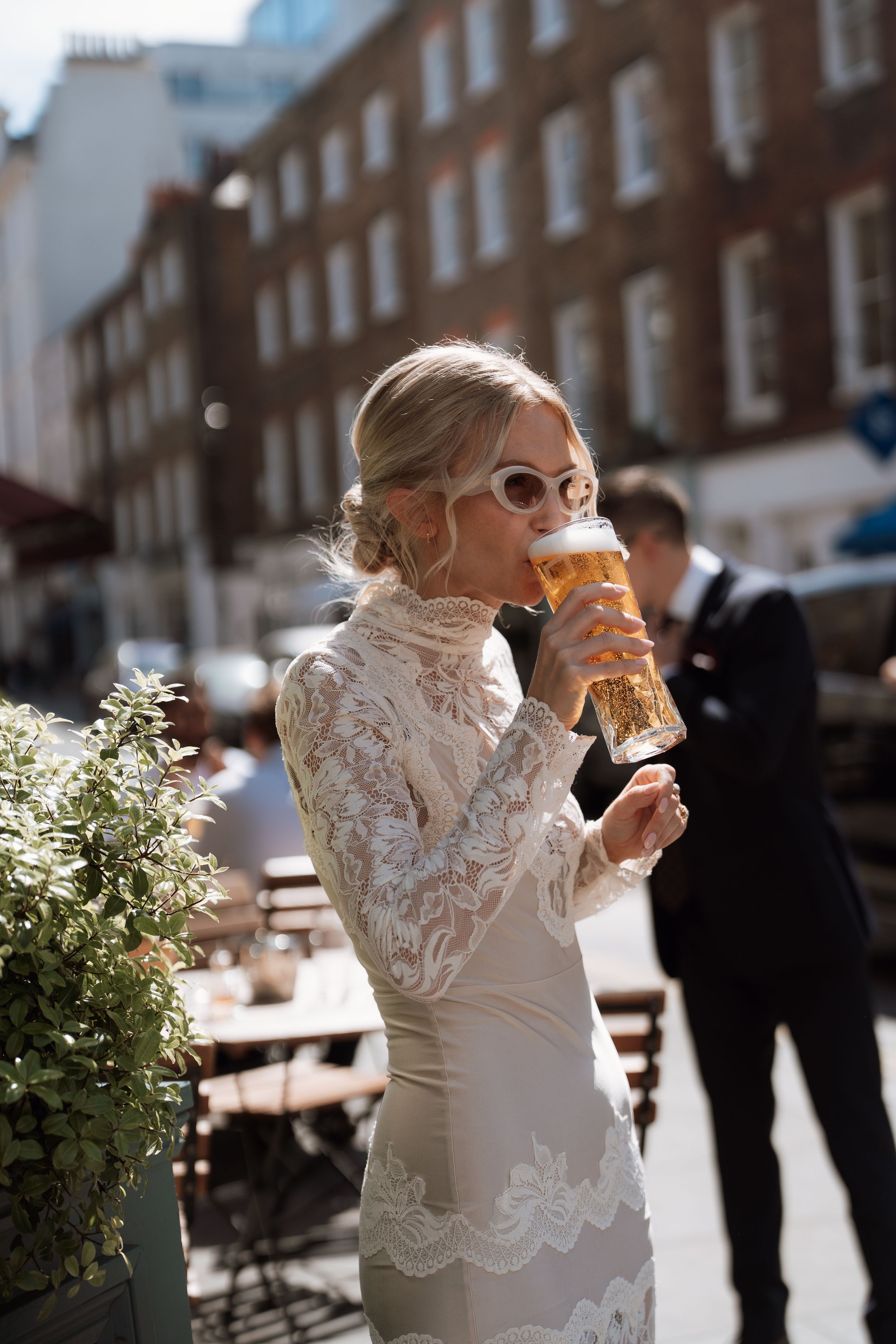 Bride in lace wedding dress and sunglasses drinking a pint of beer outdoors on a sunny city street.