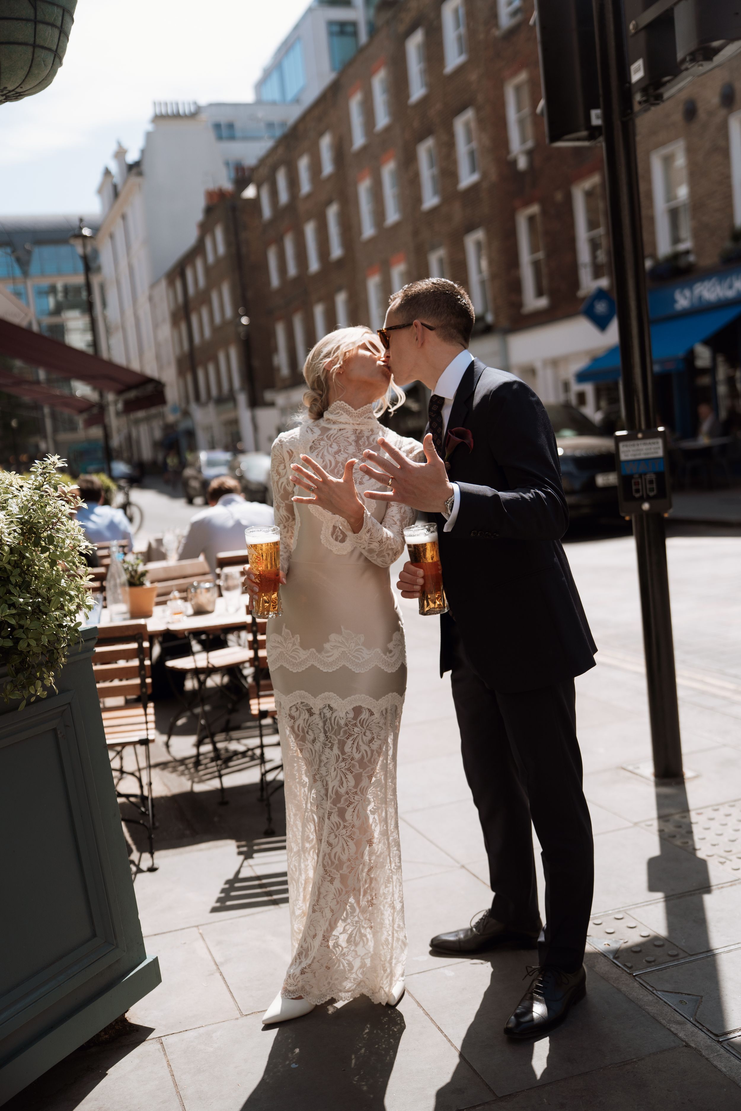 Bride in lace gown and groom in dark suit share a kiss while holding pints of beer on a sunny London street.
