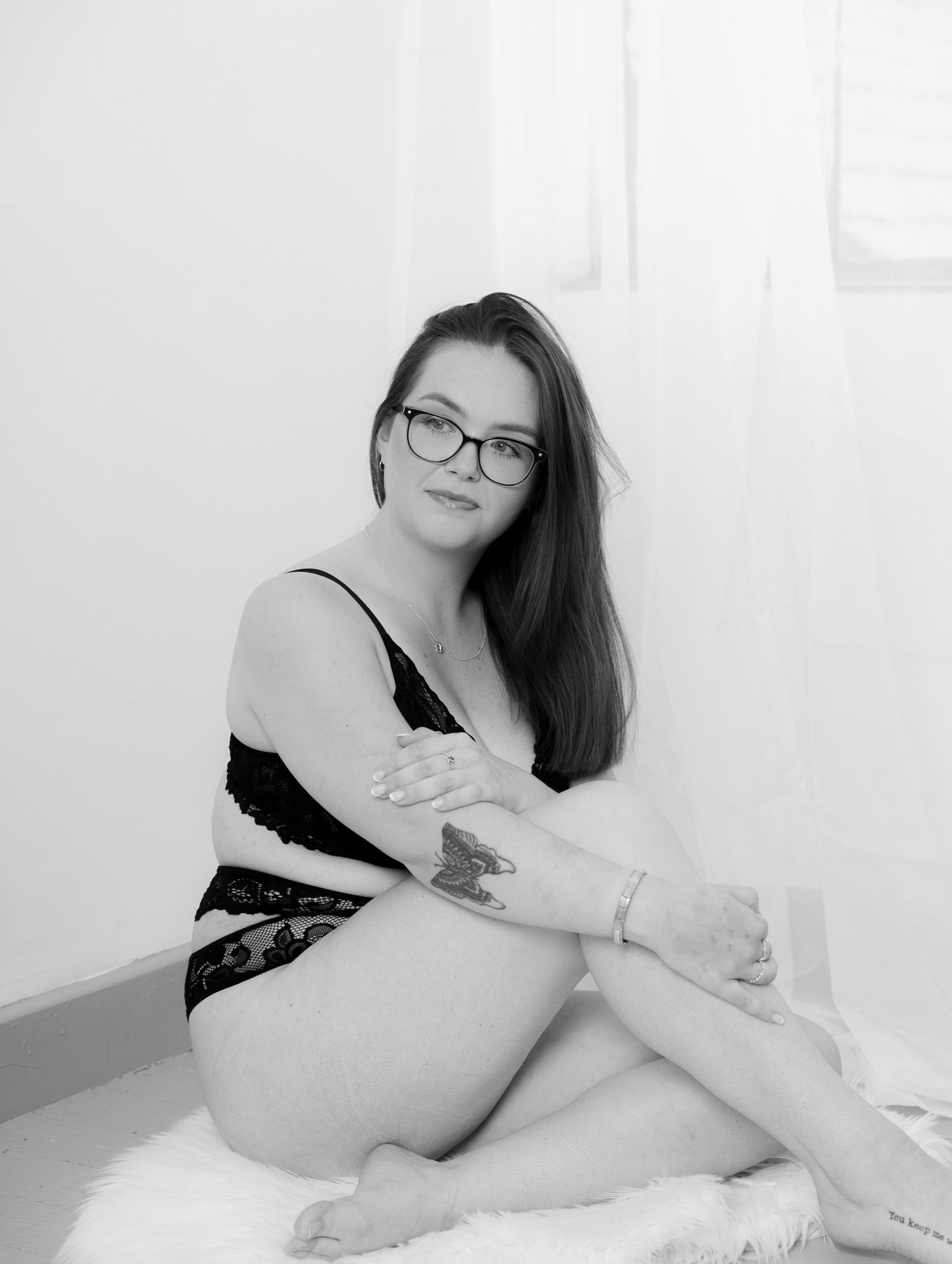 Black and white photo of a young woman with glasses and tattoos sitting in lingerie on a white surface.