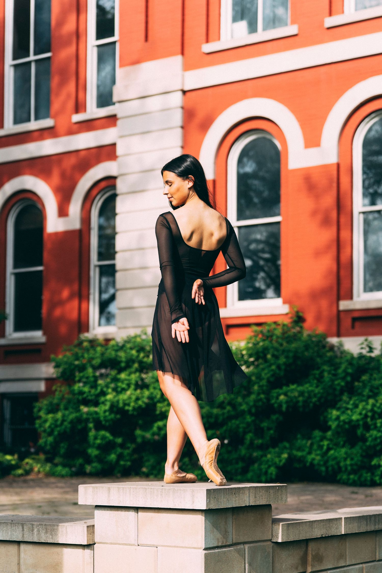 Professional senior portrait photographer captures a dancer in a sheer black dress balancing on a stone pedestal in downtown Crown Point, In