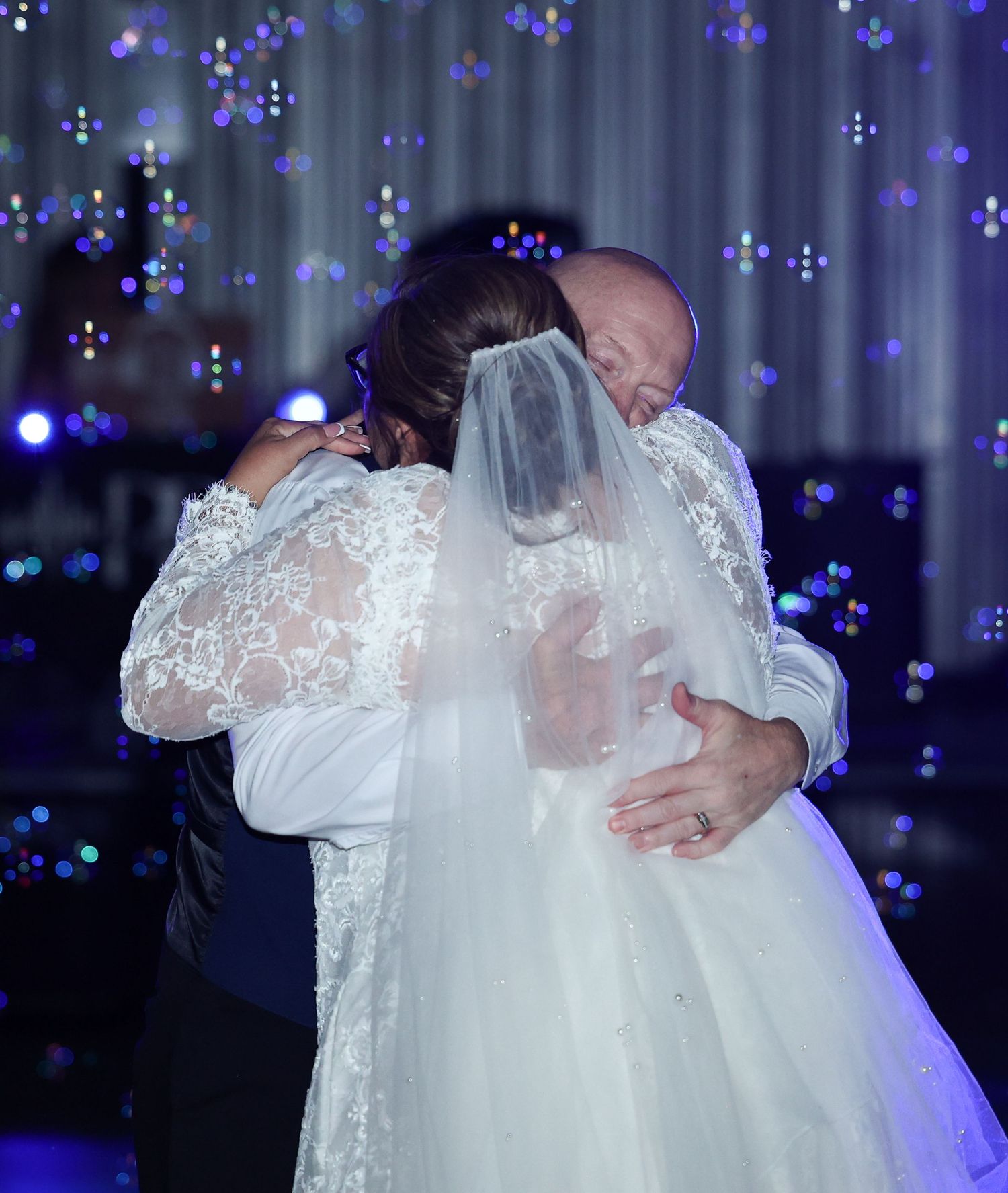 Bride and groom share emotional first dance embrace, wedding veil and lace gown, bokeh lights backdrop.