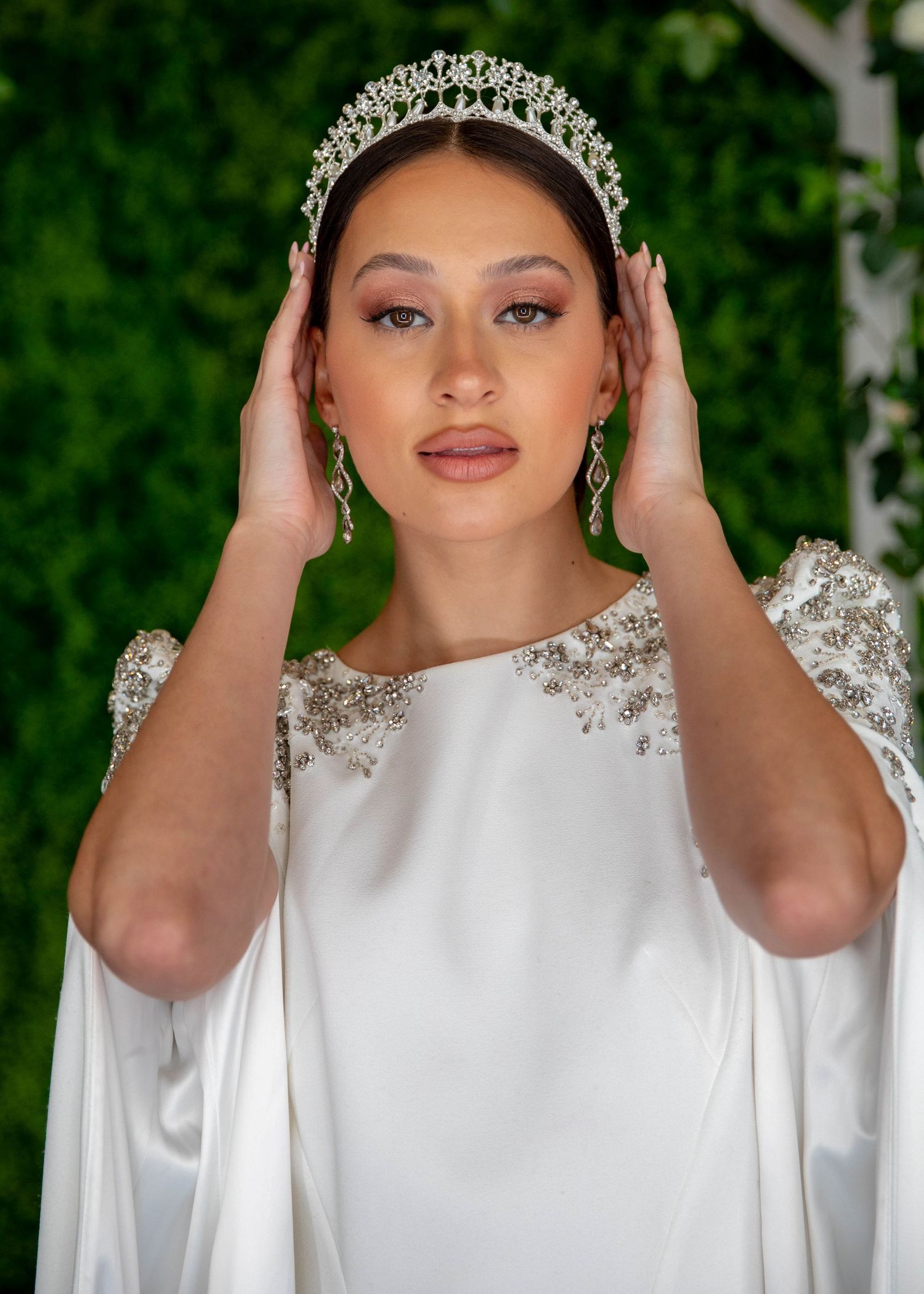 Bride wearing a crystal tiara and white beaded gown with cape sleeves, posing against a green backdrop.