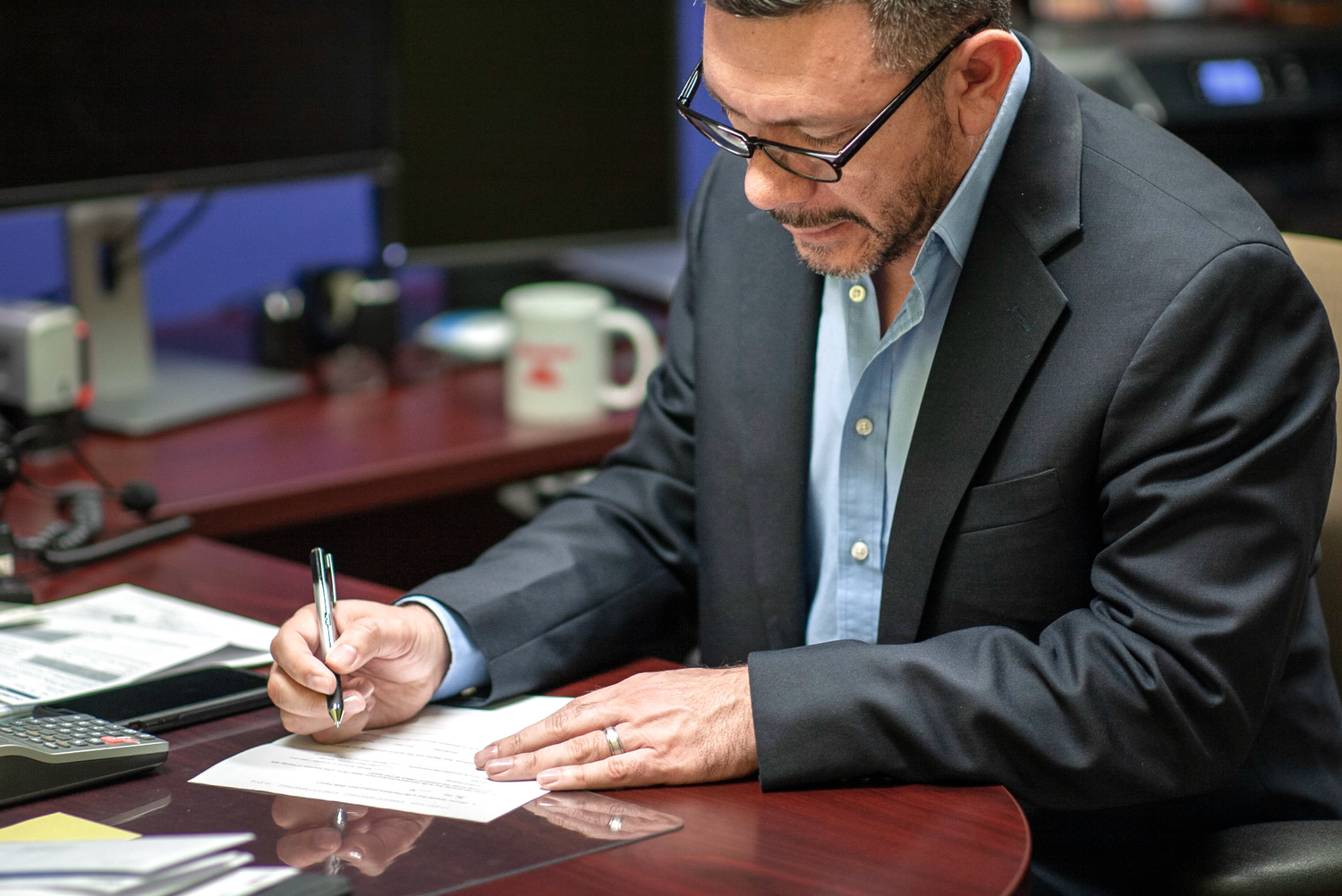 Professional businessman in suit signing documents at a mahogany desk in a modern office setting.