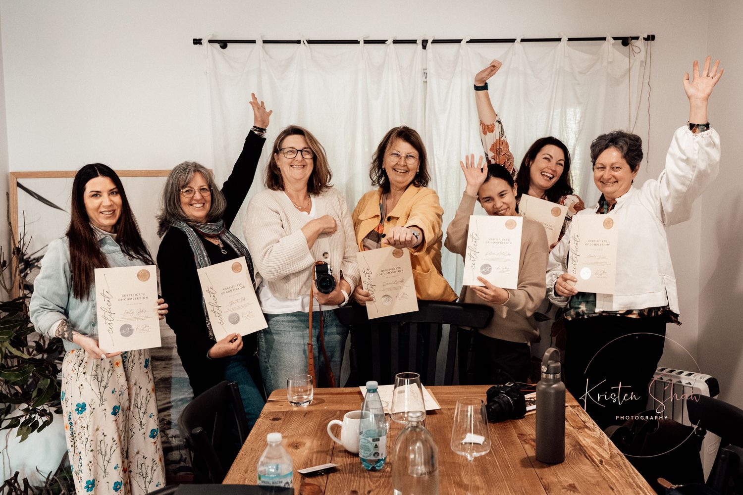 Group of seven women celebrating with certificates, raising hands joyfully around a wooden table in a bright room.