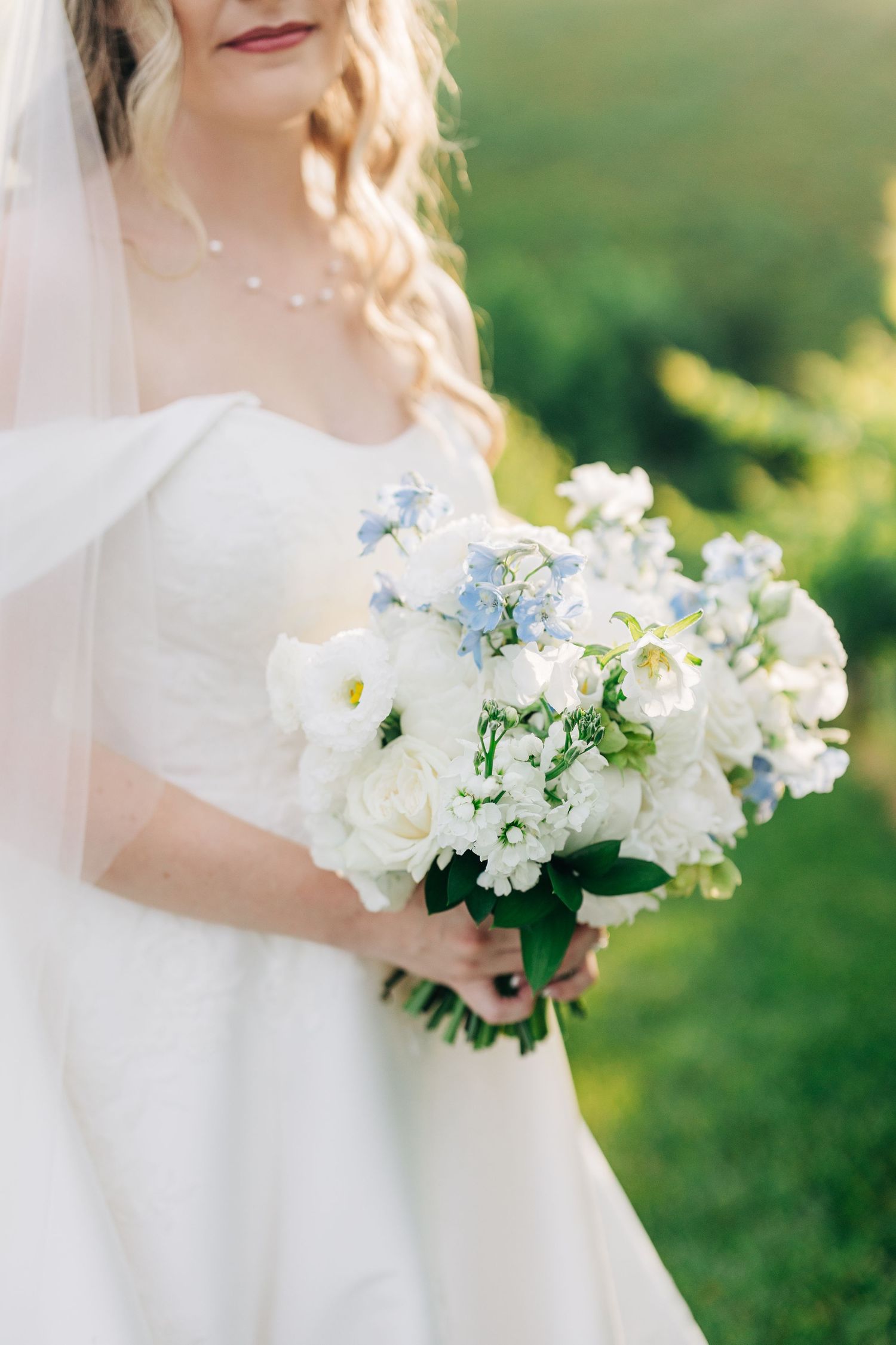 Bride in white gown holding elegant bouquet of white roses and blue flowers outdoors on her wedding day.