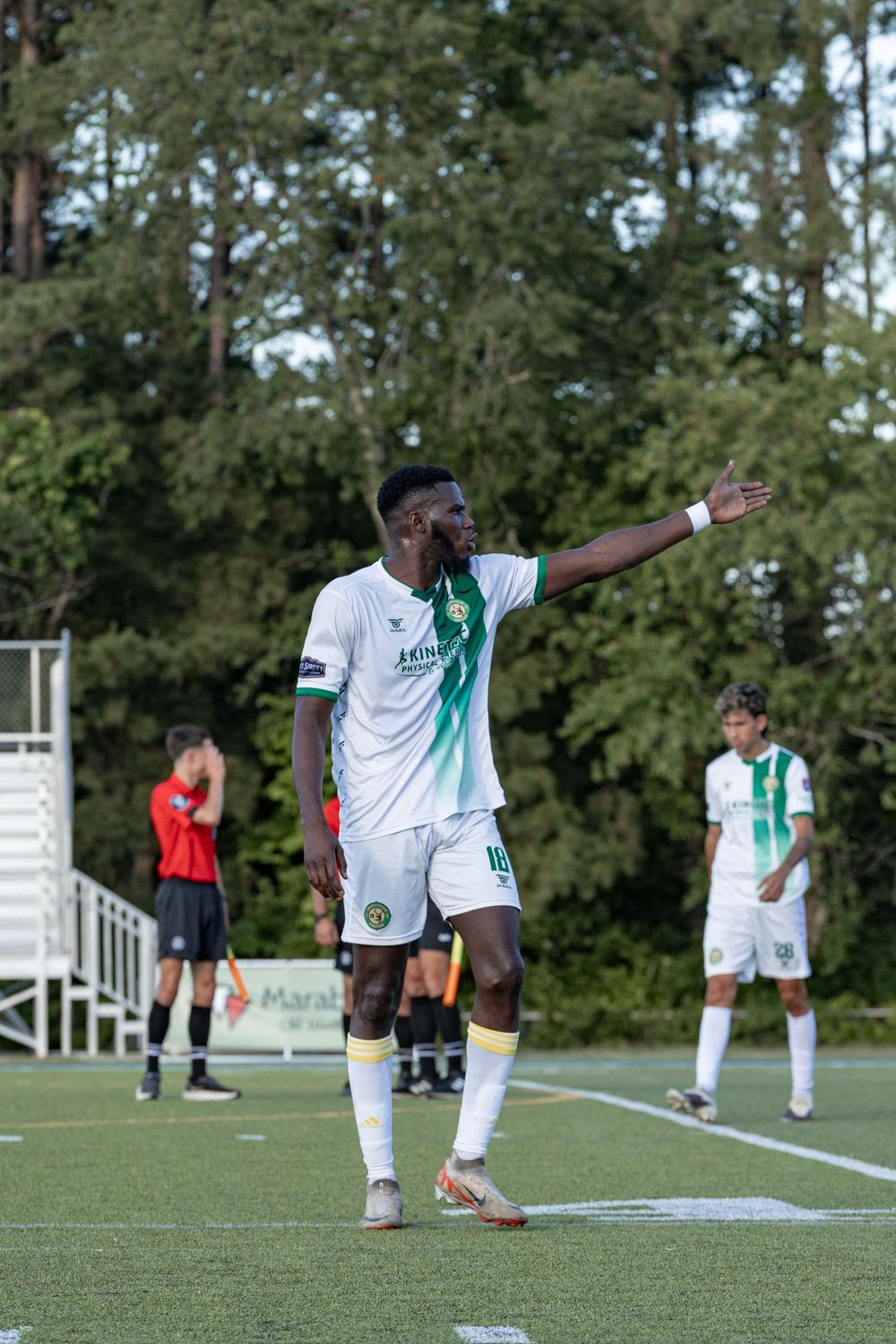 Soccer player in white uniform gestures on field during a match, with referee and teammates visible in background.
