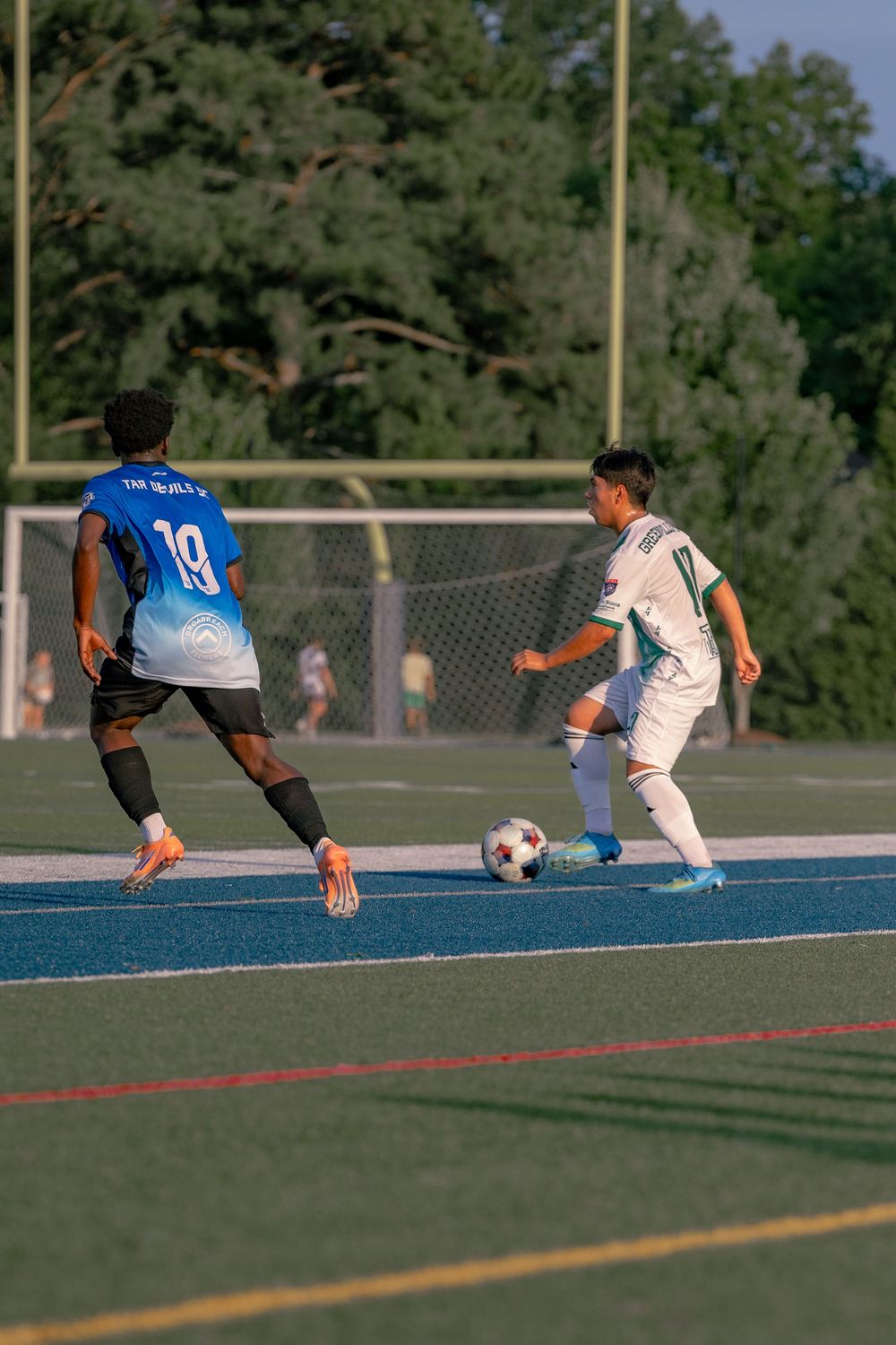 Two soccer players compete for the ball on a blue turf field, with player 19 in blue defending against a white-uniformed opponent.