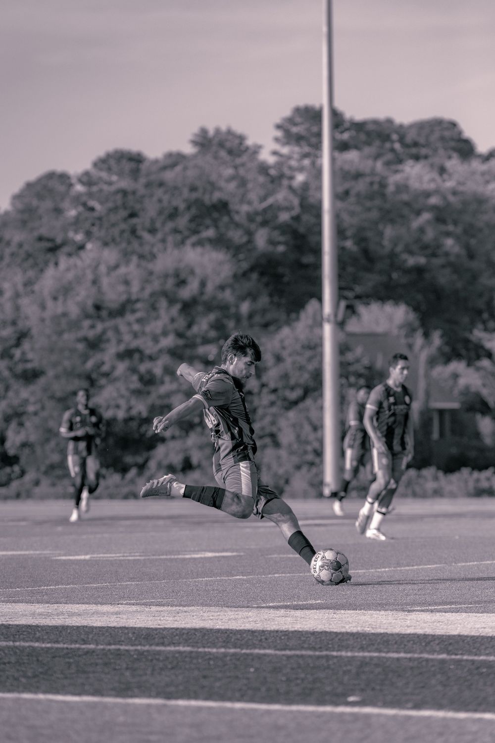 Black and white photo of a soccer player mid-kick on a field with teammates and trees in the background.