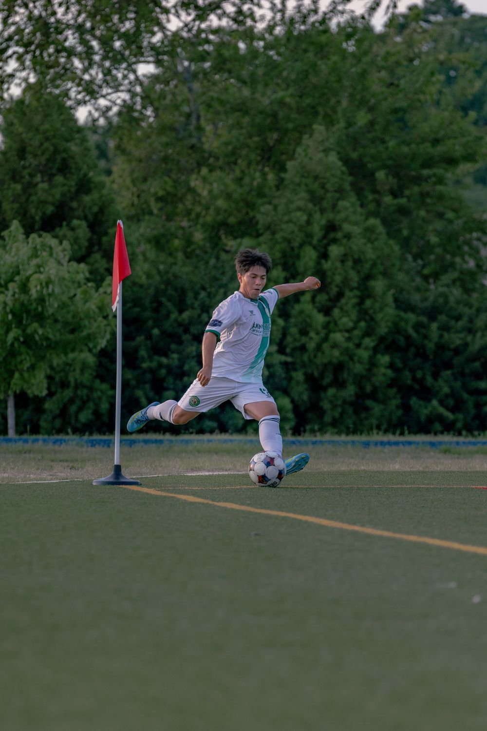 A soccer player in white kit takes a corner kick near a red flag during an outdoor evening game on a grass field.