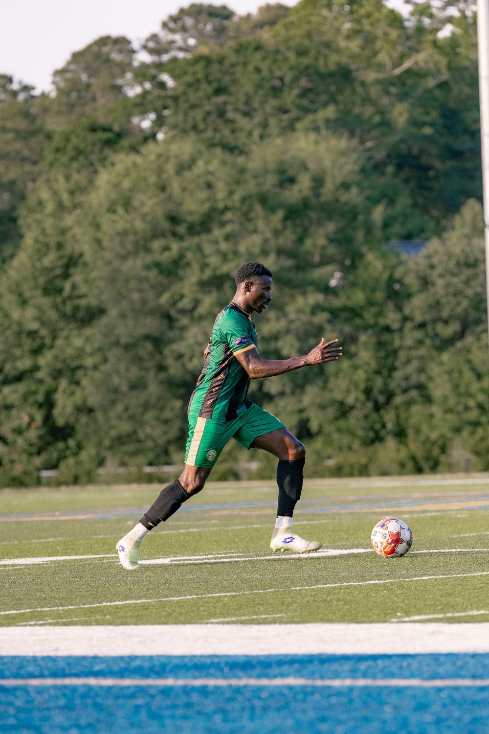 Soccer player in green uniform dribbling ball on turf field with trees in background.