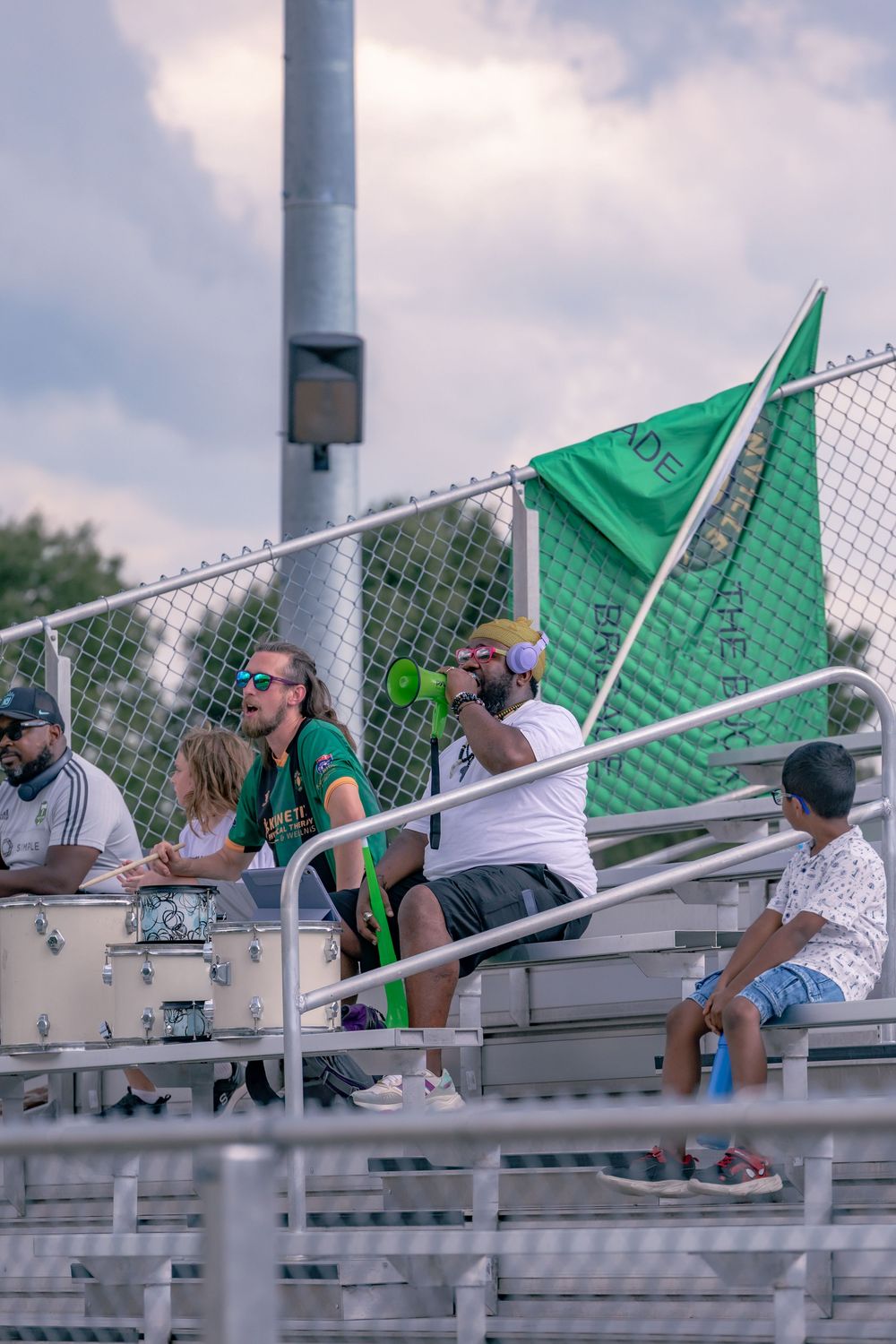 Sports fans cheer in bleachers with a green flag, one blowing a megaphone at an outdoor stadium event.