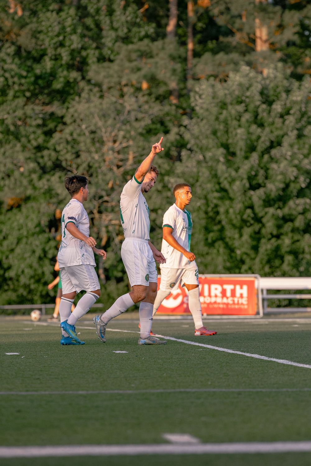 Soccer player raises finger in celebration on field, teammates nearby, lush green trees in background during evening match.
