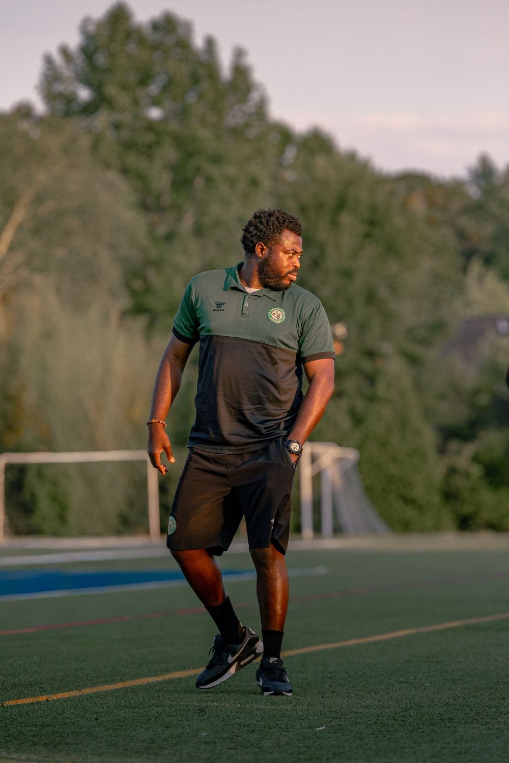 A man in a green and black polo shirt and shorts walks on a sports field surrounded by trees.
