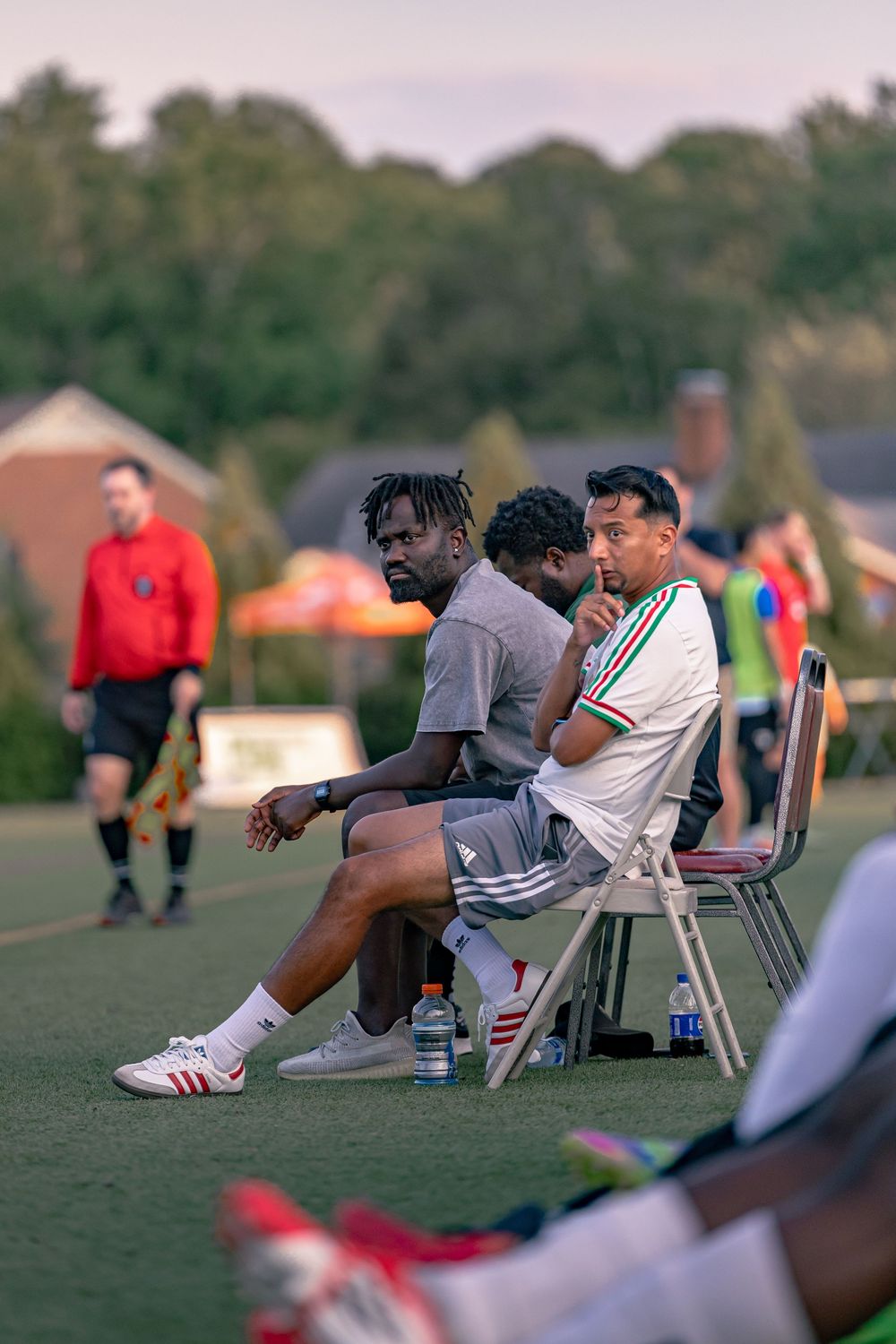 Soccer players and coaches sit on the sideline bench during an outdoor match at dusk, with a referee visible in background.