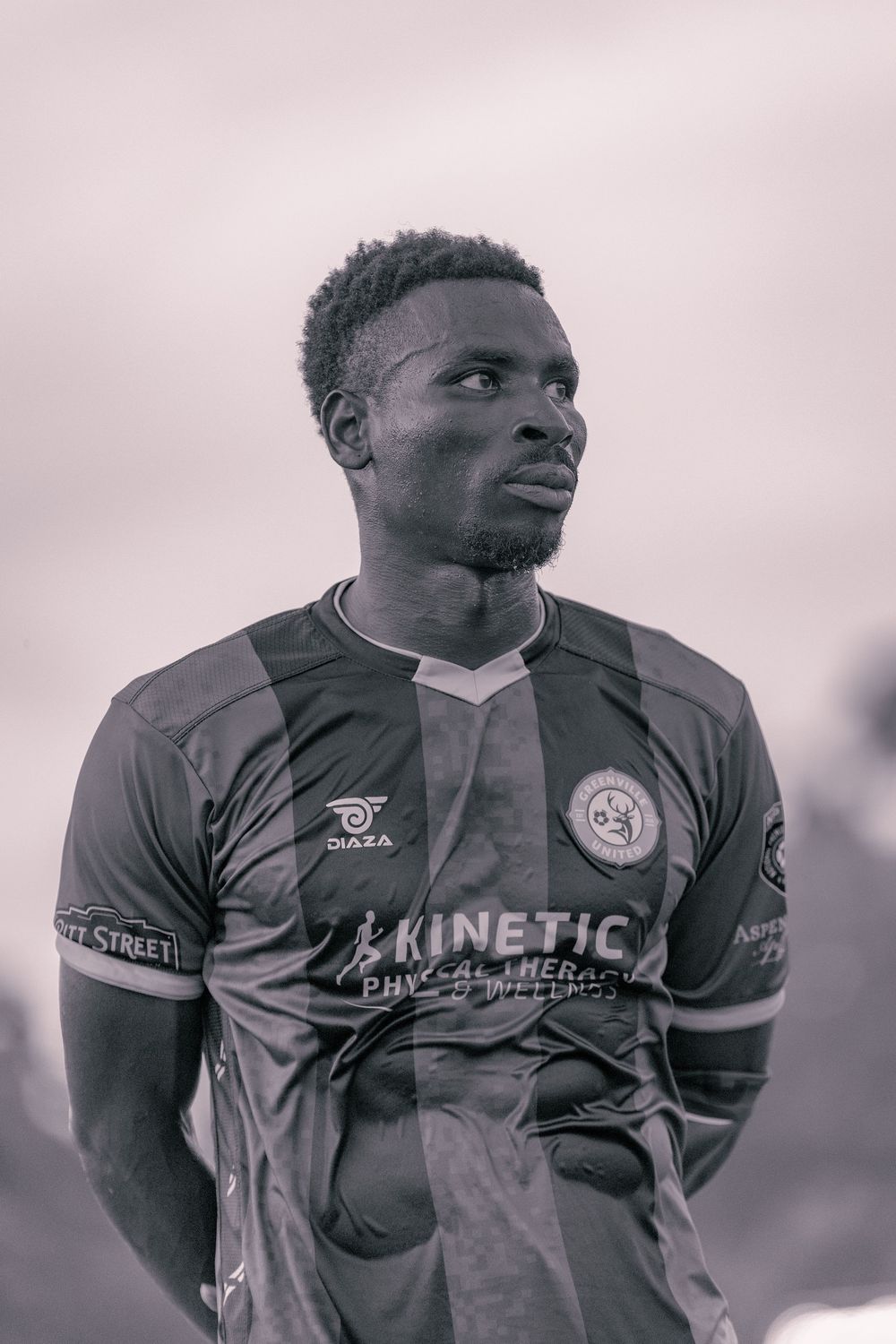 Black and white portrait of a male soccer player wearing a Kinetic Physical Wellness jersey on a cloudy day.