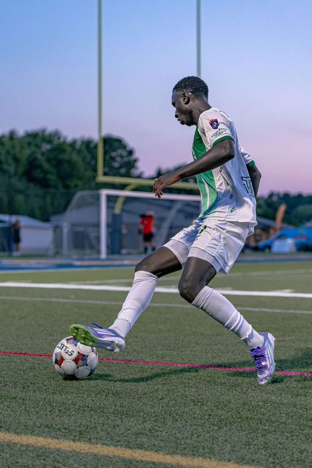 Young soccer player in white and green uniform dribbling ball on turf field at dusk.
