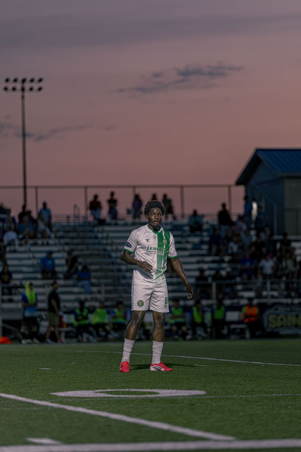 Soccer player in white and green uniform stands on field at dusk with stadium crowd in background.