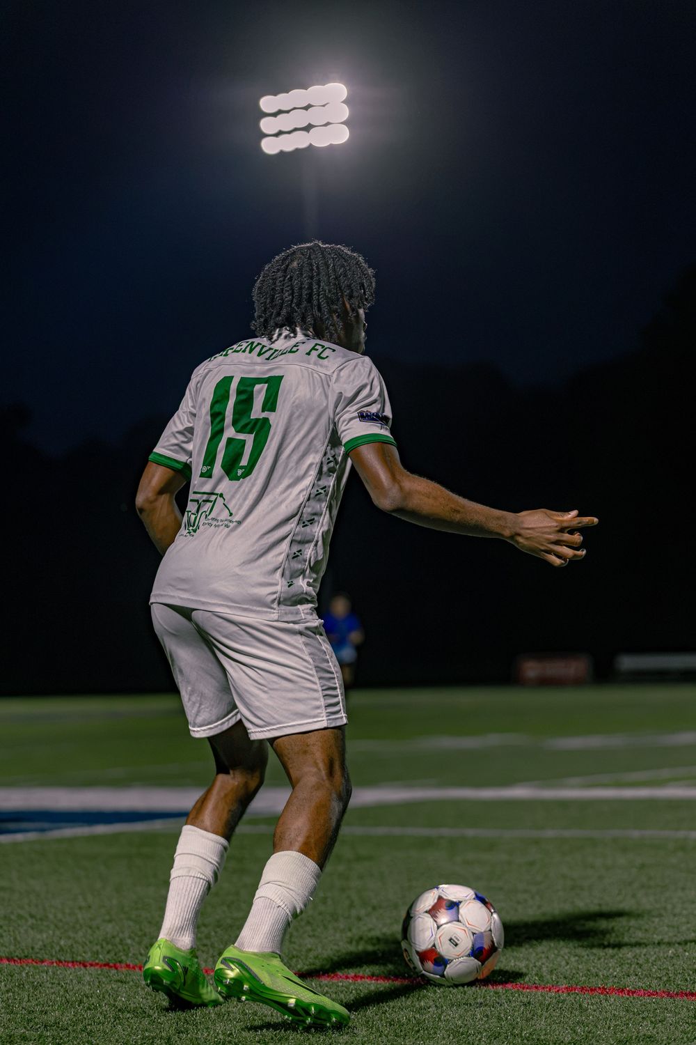 Soccer player #15 in white and green uniform dribbles ball under stadium lights at night game.
