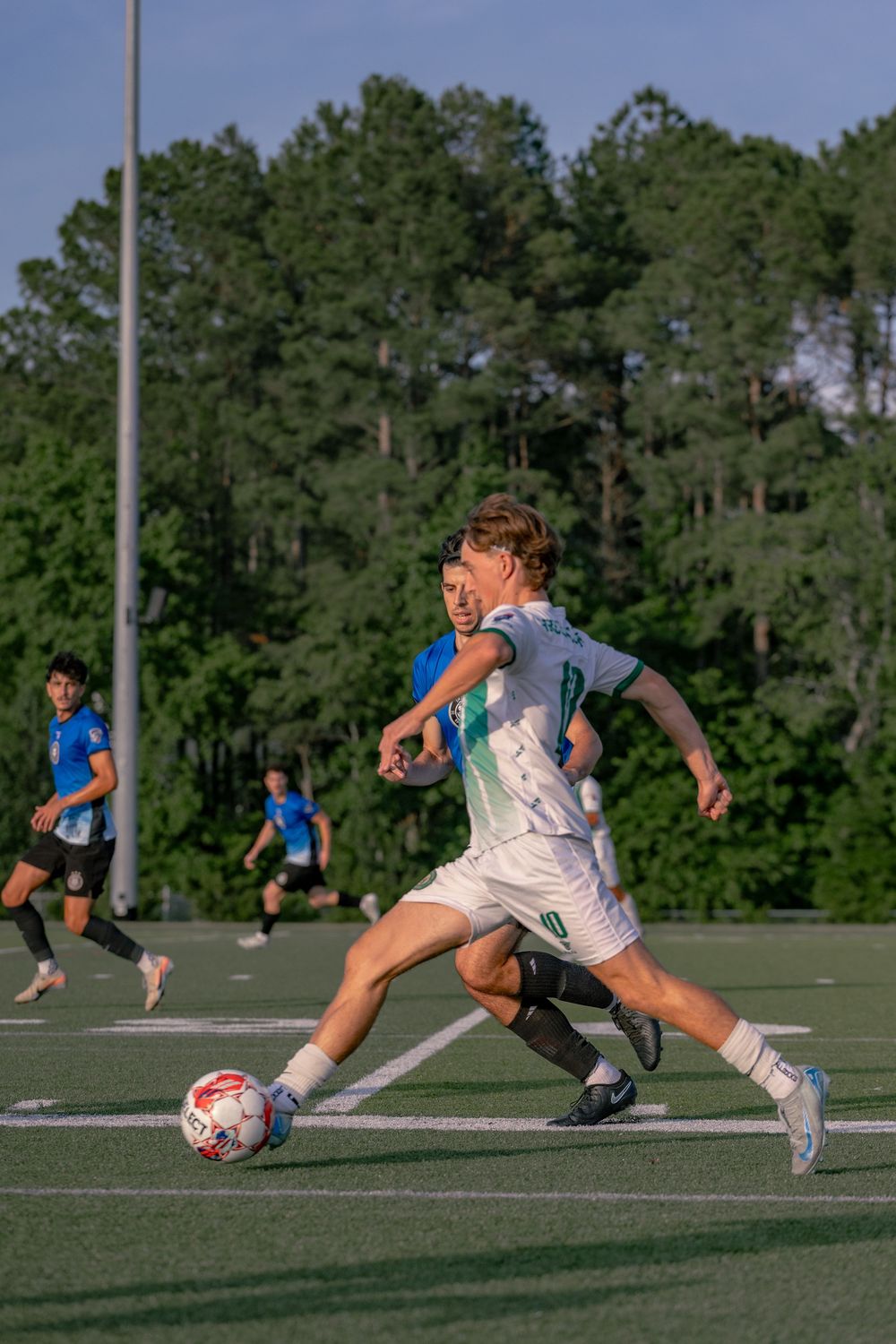 A young soccer player in white and green uniform dribbles the ball during an evening match on a turf field.