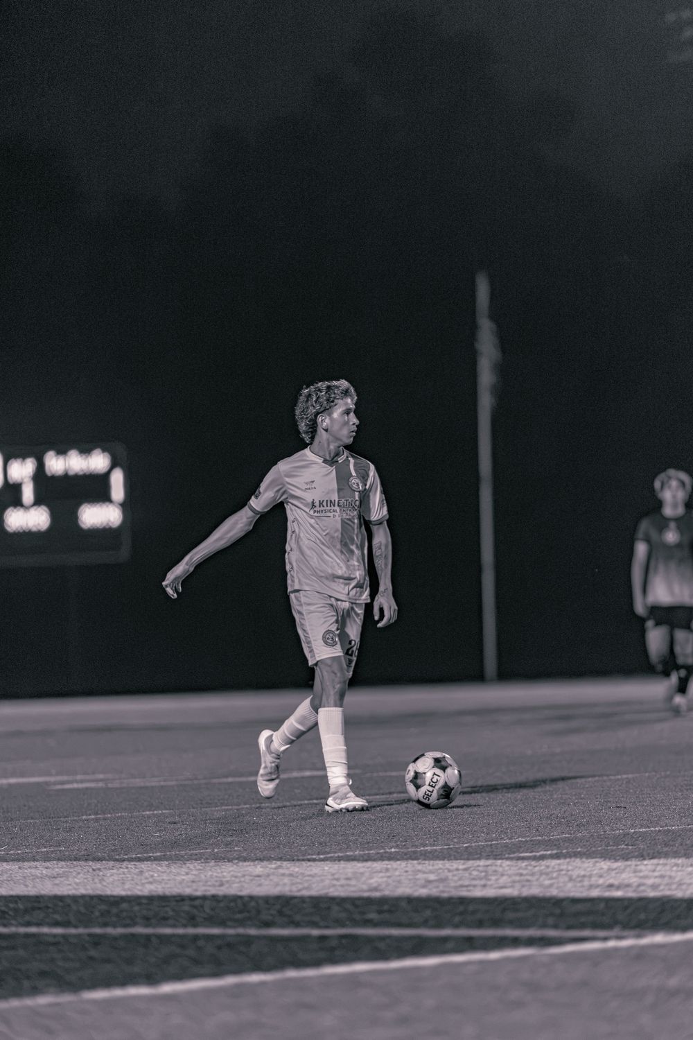 Black and white photo of a soccer player dribbling a ball on a field at night under stadium lights.