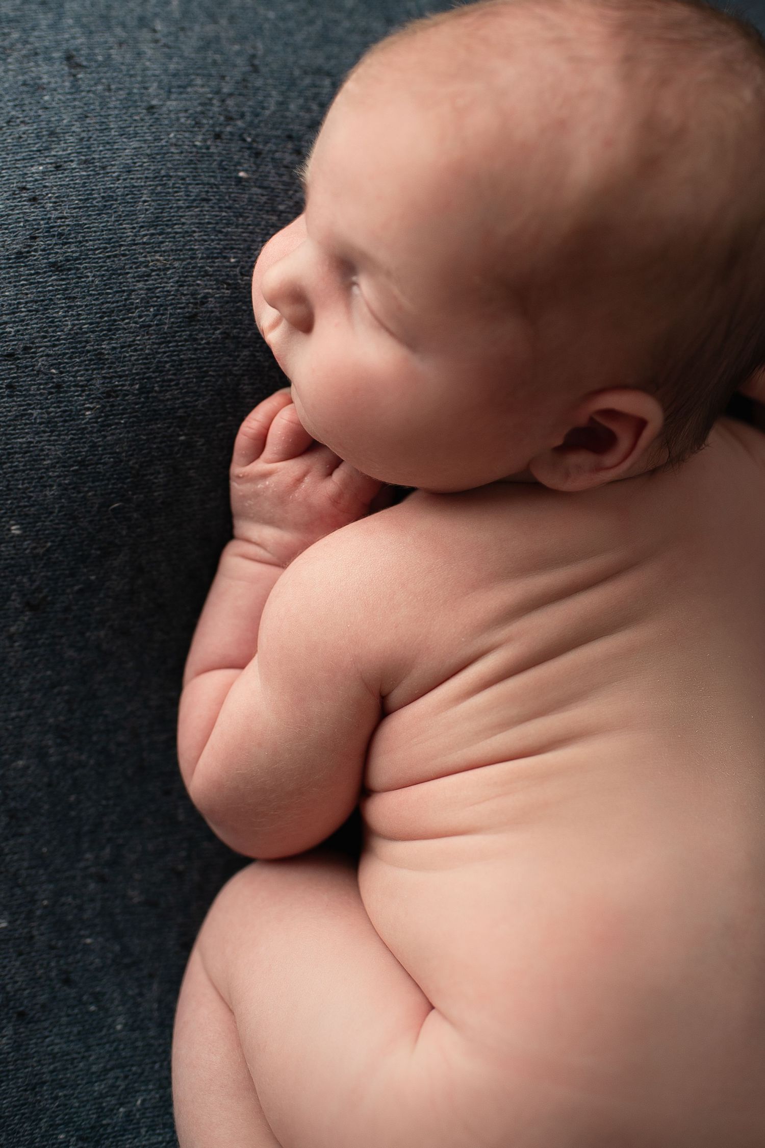Sleeping newborn baby curled up on a dark gray textured blanket.