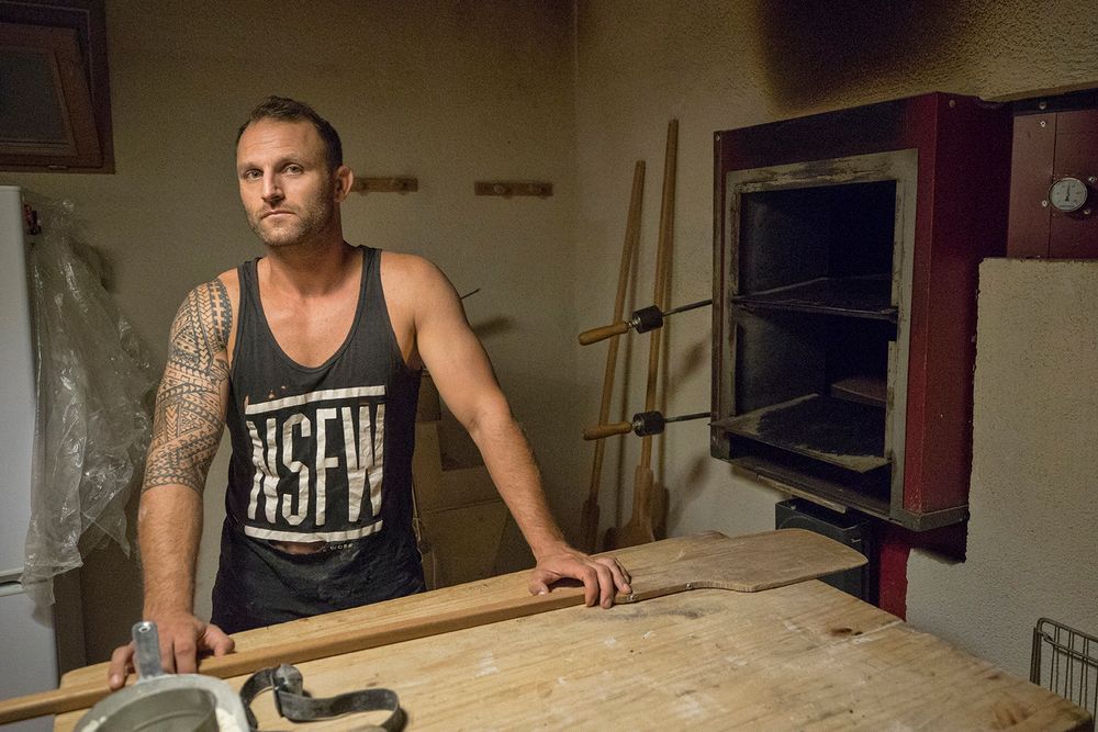 Tattooed man in NSFW tank top standing in front of a large oven in a bakery.