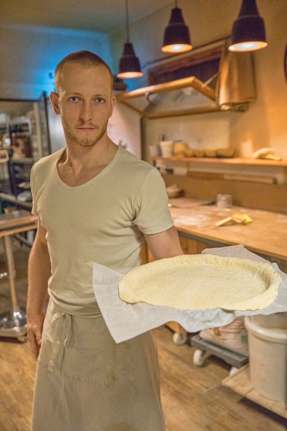 Baker holding raw pie crust on parchment paper in a warmly lit professional kitchen.
