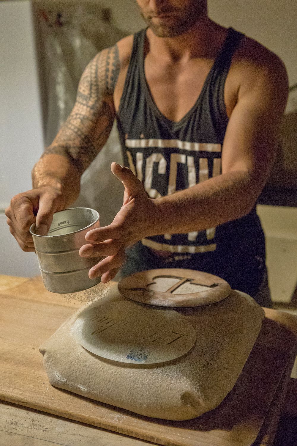 Tattooed male baker weighing dough on a scale in a rustic bakery workshop.