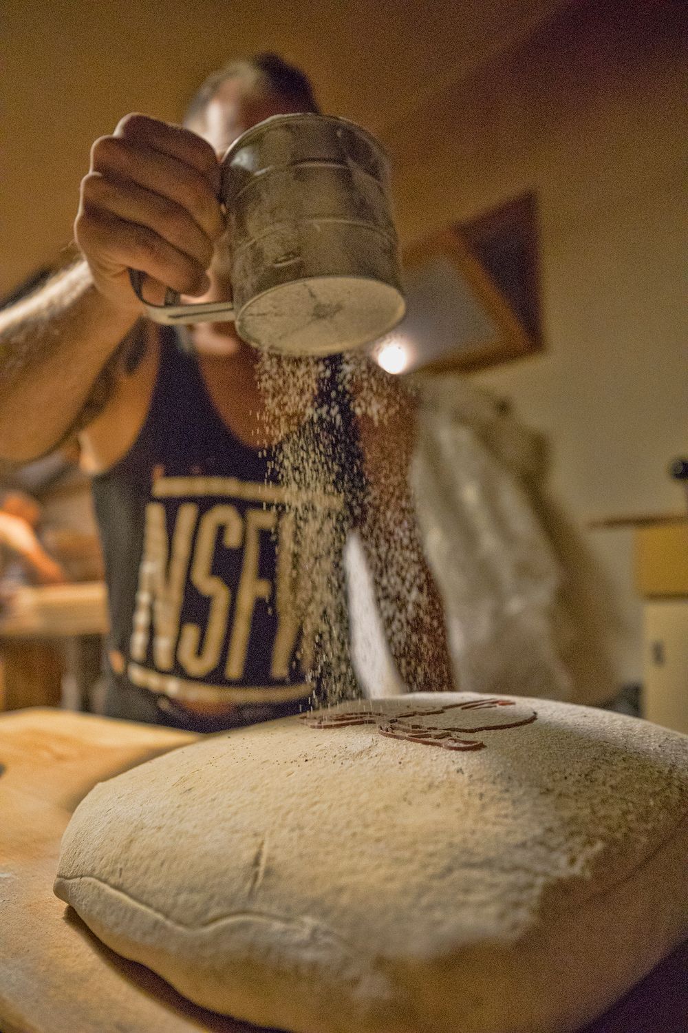 Baker sifting flour over shaped bread dough on a wooden work surface.