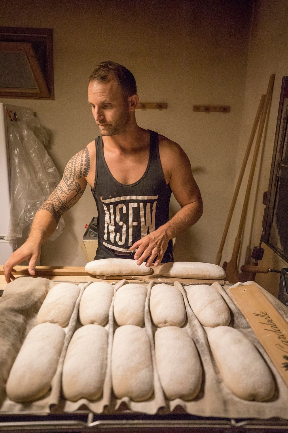 Tattooed baker arranging rows of shaped bread loaves on a proofing cloth.