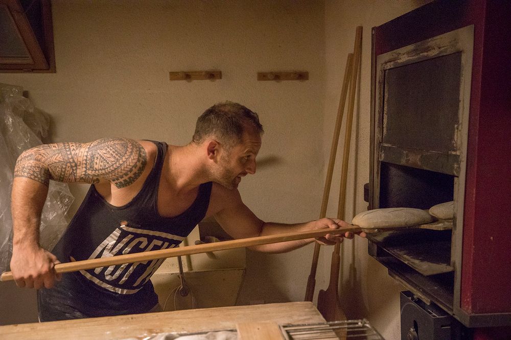 Male baker loading bread into a stone oven using a long wooden peel.